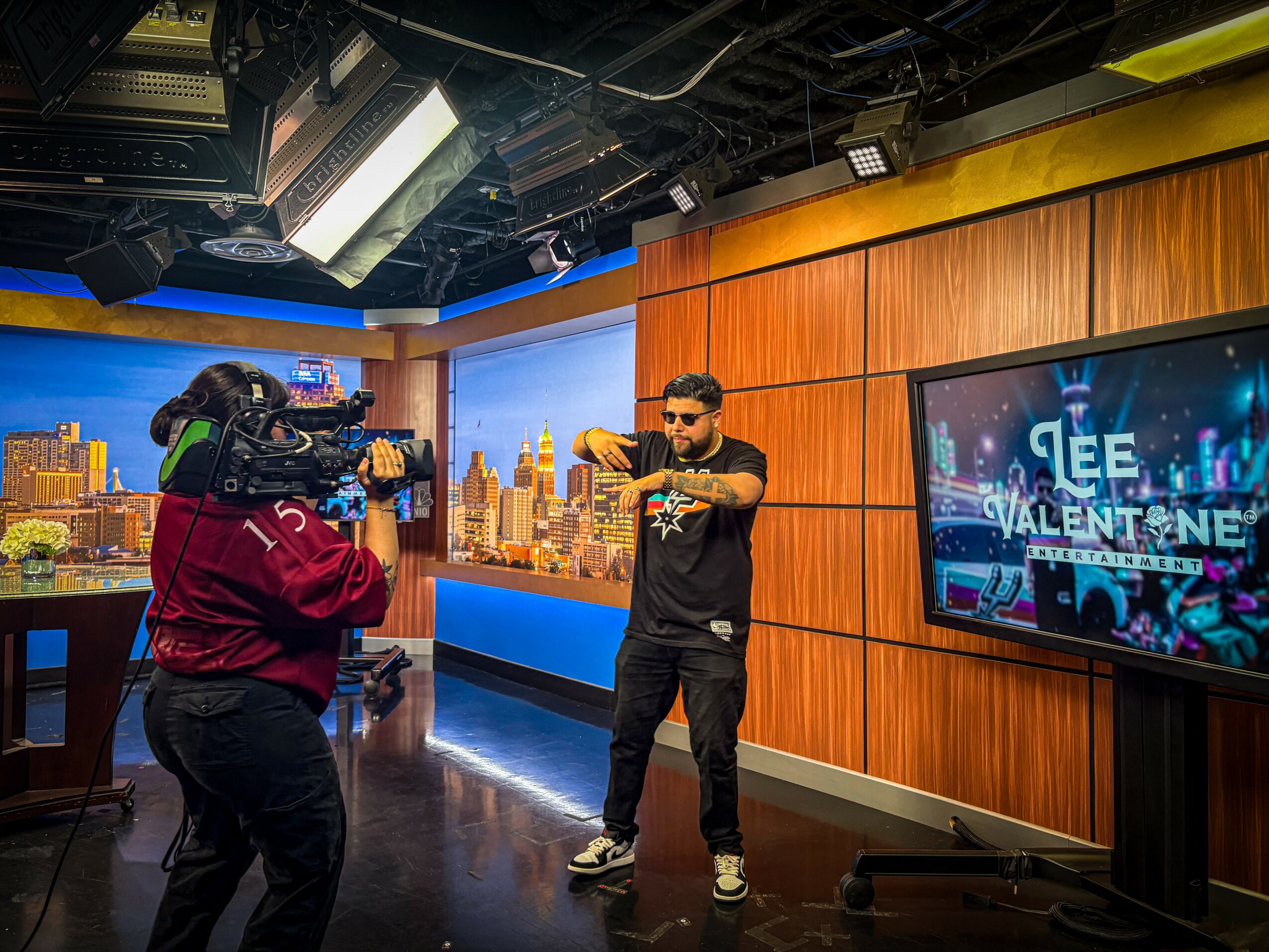 Person in black clothing and sunglasses pointing at camera, studio with cityscape background and TV screen displaying 'Valentine' text.