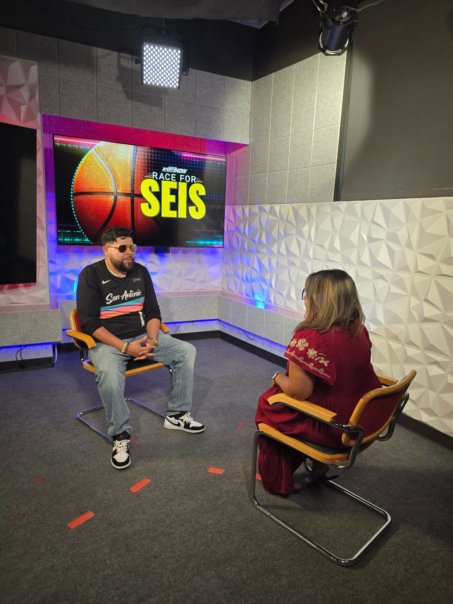 Person sitting on a chair with a backpack, facing a woman with a backpack, in front of a colorful sign that says 'SIES'.