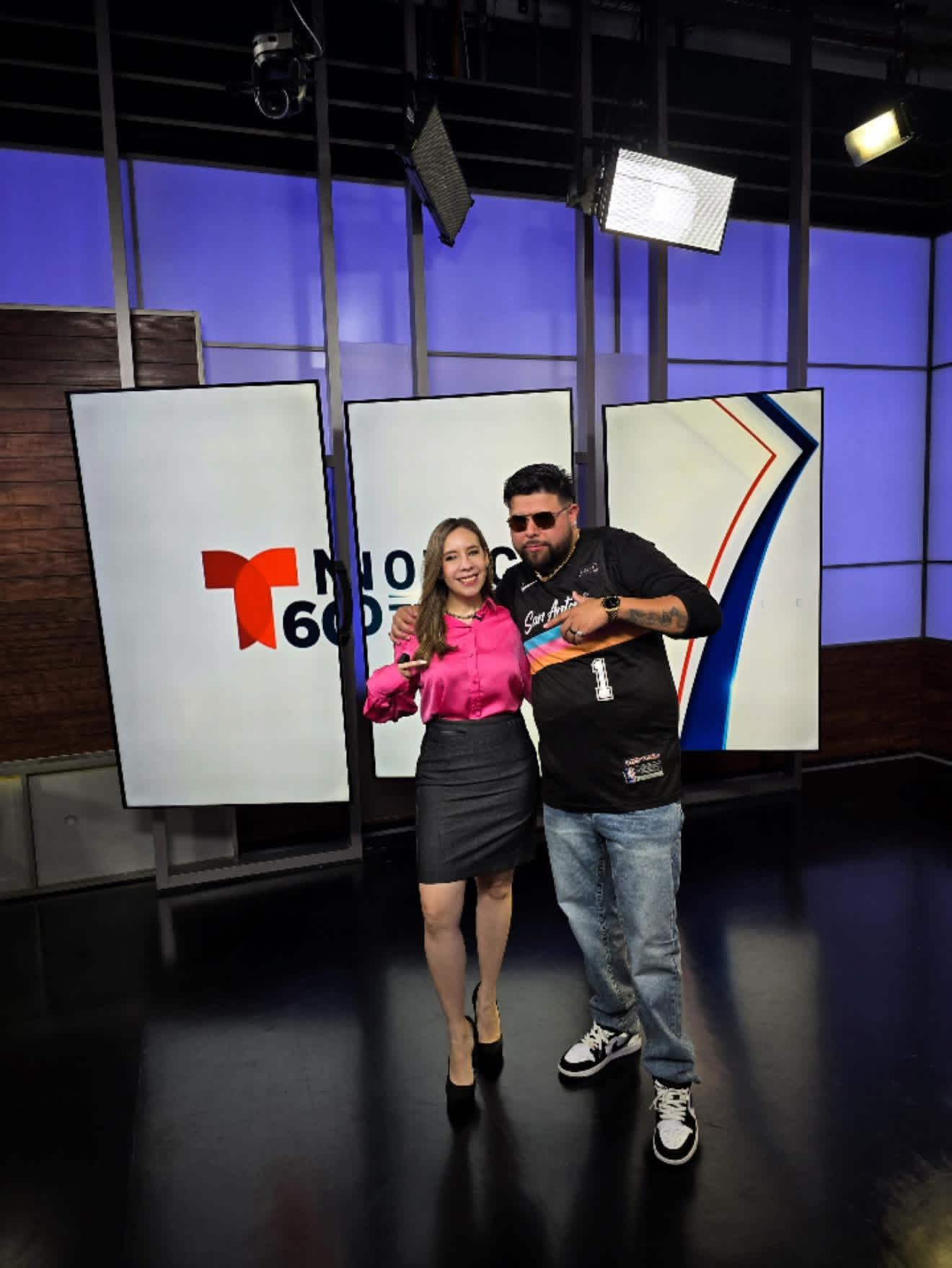 Two people standing in a TV studio with large screens and studio lights behind them.