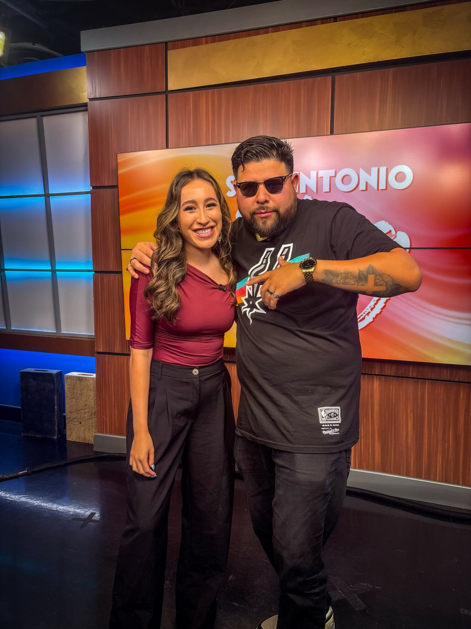 A woman and a man pose together in a studio, smiling, with a colorful background and a screen displaying the name Antonio.