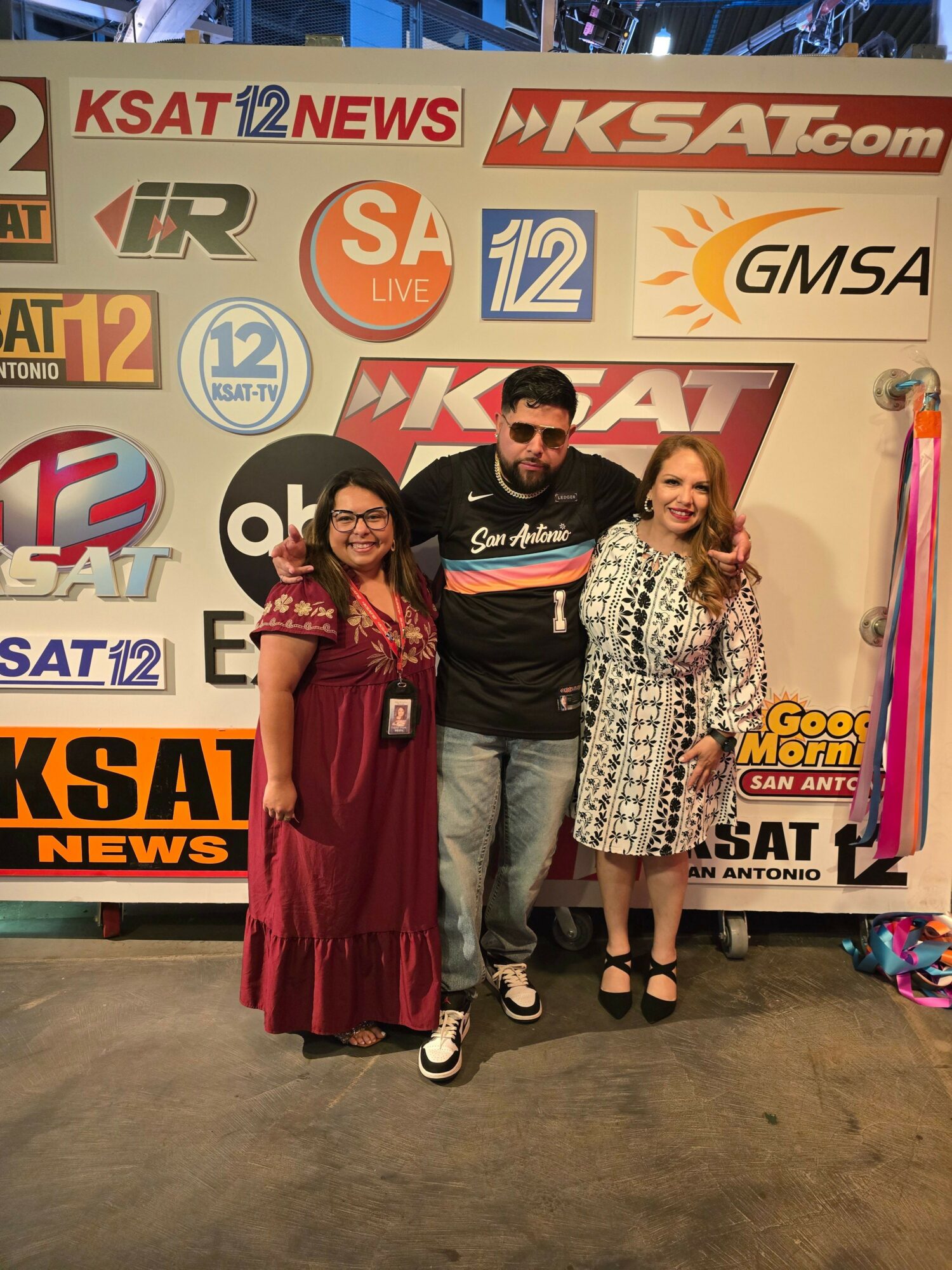 Three women standing in front of a news station backdrop, smiling, with various logos and text behind them.