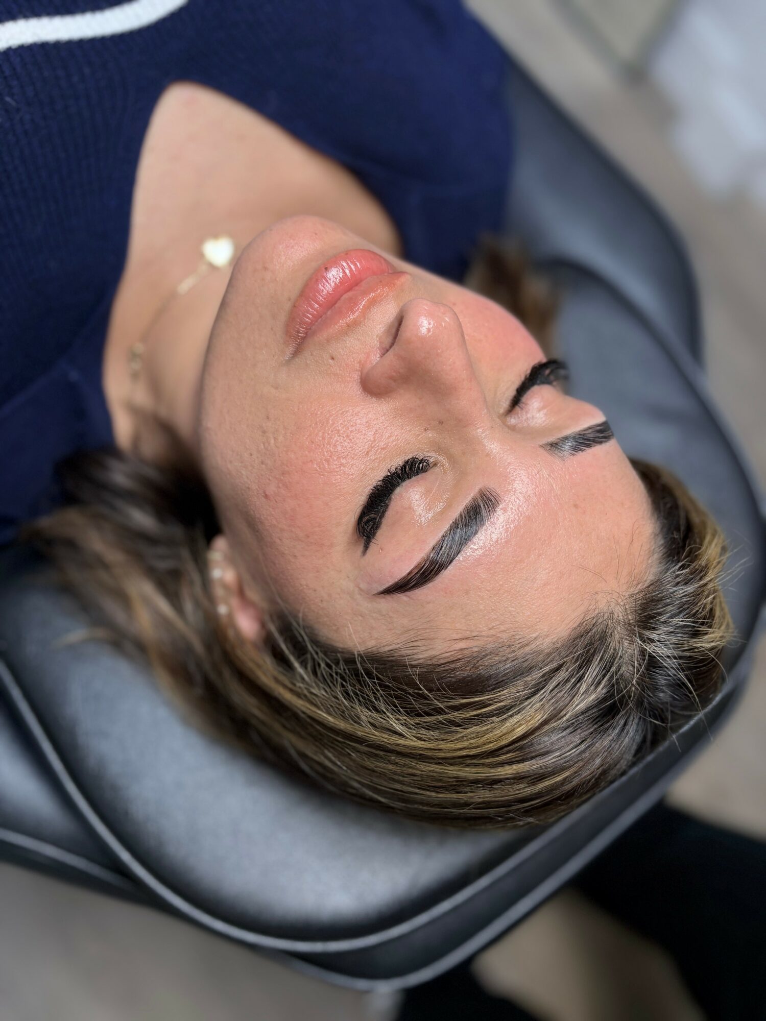 Woman with closed eyes lying on a medical chair, with makeup and a necklace, in a clinical setting.
