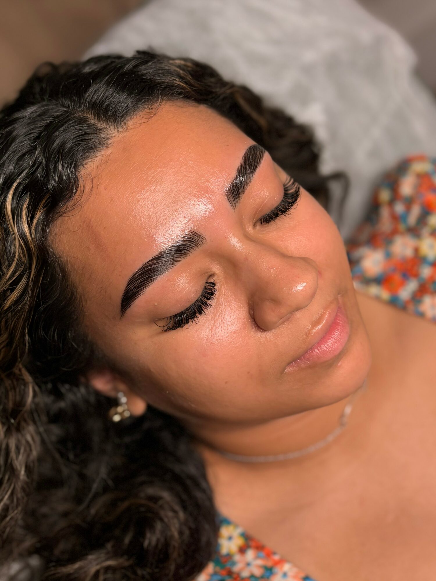 Close-up of a woman with dark curly hair, closed eyes, and makeup, lying down on a bed with a patterned pillow.
