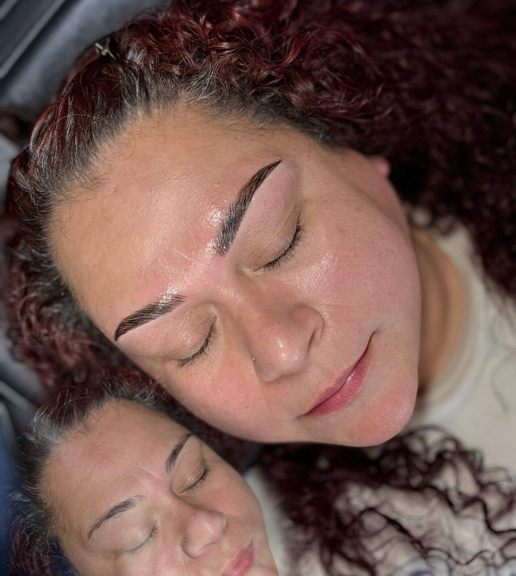 Two women with closed eyes, one with curly hair and the other with straight hair, resting their heads together.