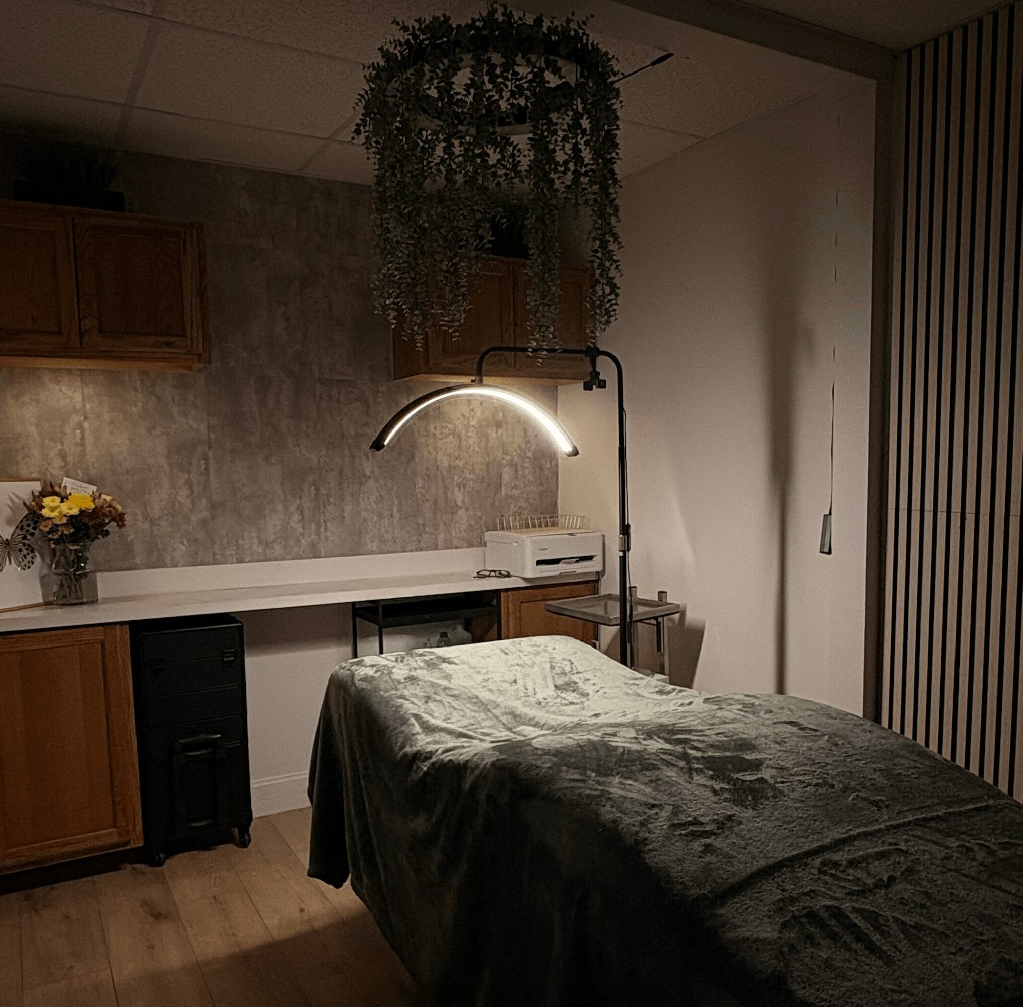 Massage table covered with dark sheet in a room with hanging plant, lamp, and wooden furniture.