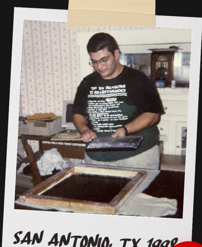 Person with glasses wearing a black T-shirt, holding a book or folder, standing in a room with patterned wallpaper and furniture.