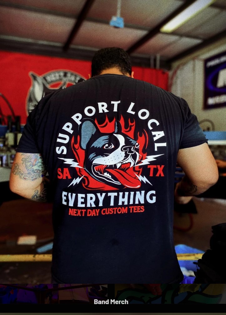 Person wearing a black T-shirt with a graphic of a raccoon, flames, and text, standing indoors with a red background.
