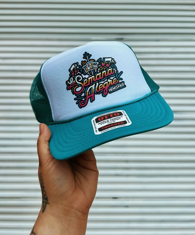 Hand holding a teal and white trucker hat with colorful embroidered text and graphics, against a striped background.