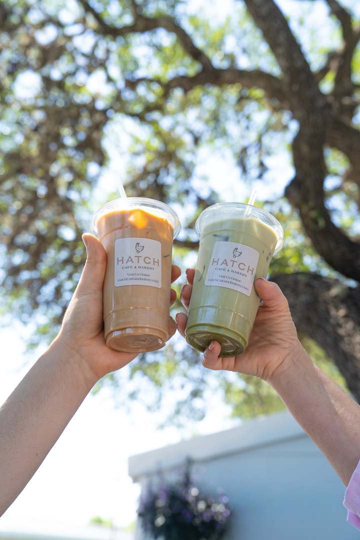 Two hands holding iced drinks with straws, one brown and one green, against a background of trees and sky.