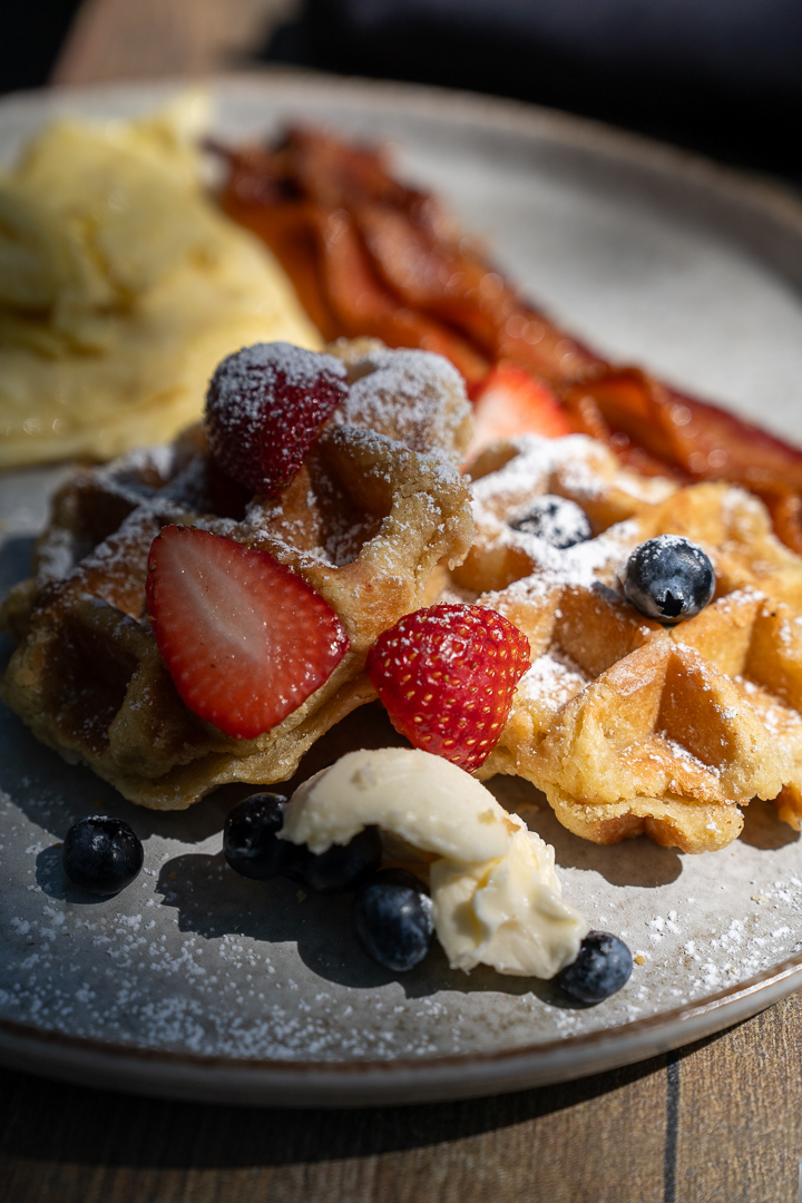 Dessert with strawberries, blueberries, whipped cream, and waffle pieces on a plate, with bacon and mashed potatoes in the background.