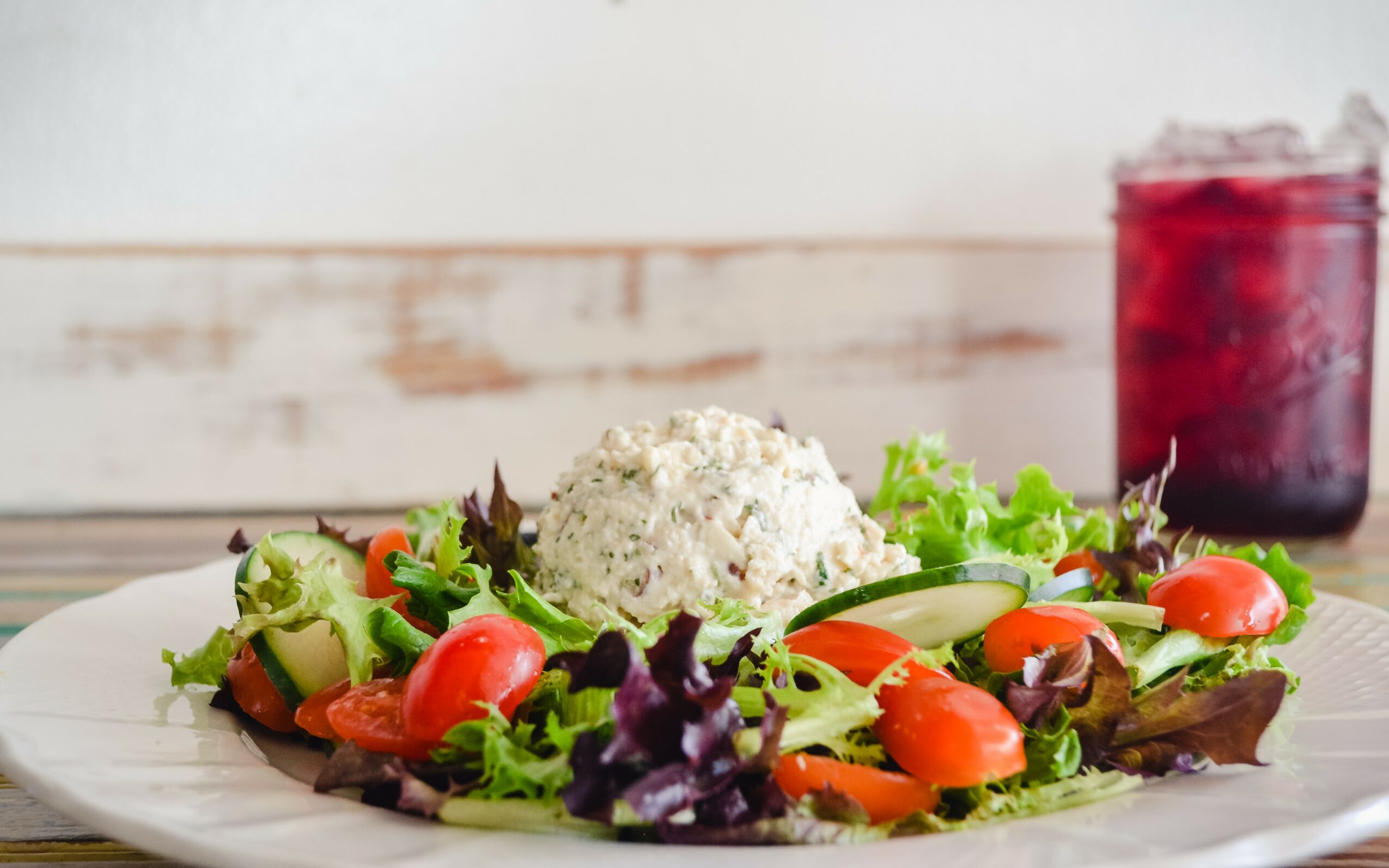 Salad with mixed greens, cherry tomatoes, and a scoop of white cheese, with a glass of red beverage in background.