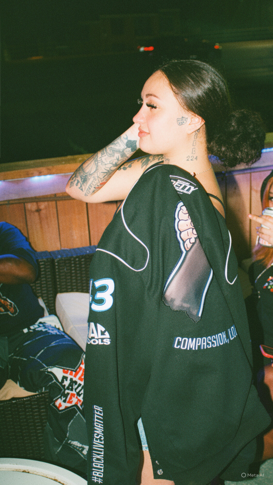 Young woman with dark hair and tattoos, sitting outdoors at night, wearing a black jacket with white text, holding a phone.