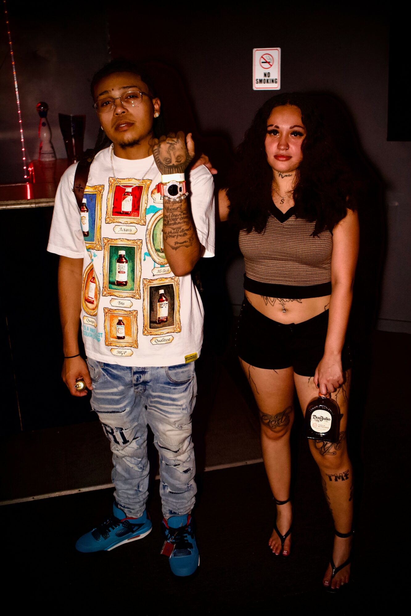 Two people standing indoors, one with glasses and a colorful shirt, the other with dark hair and a crop top, holding a bag.