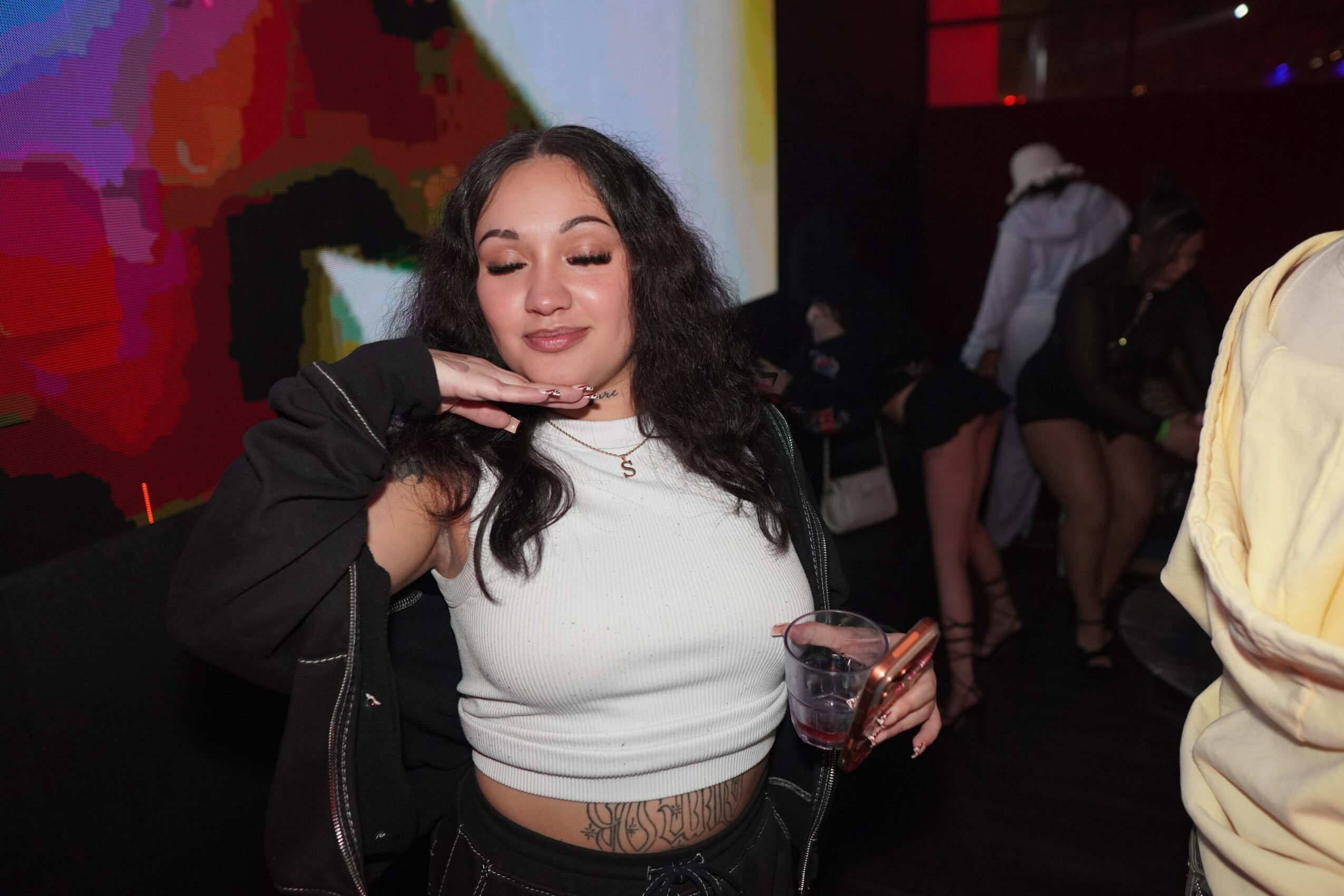 Young woman with long dark hair holding a drink, smiling with eyes closed, in a dimly lit social setting.