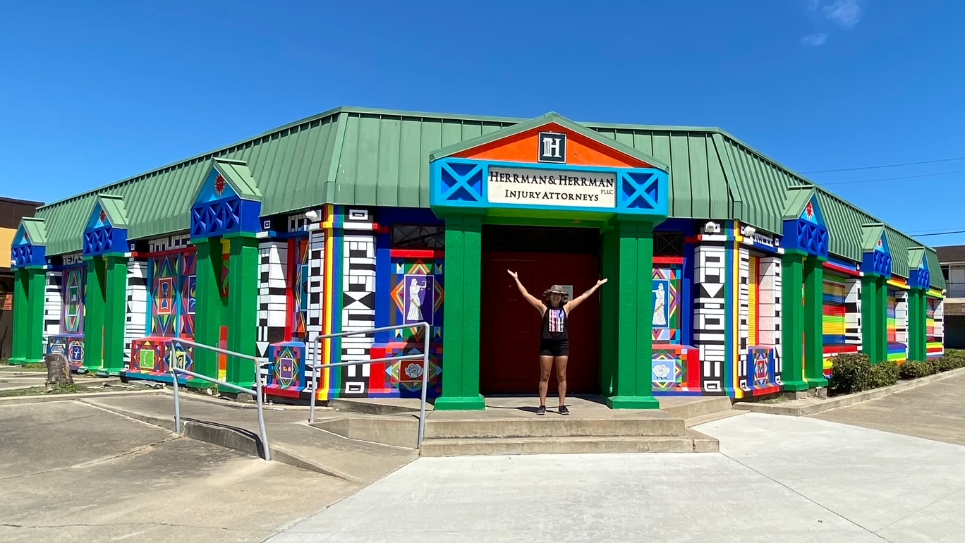 Person standing with arms outstretched in front of a colorful building with a green roof and vibrant murals.