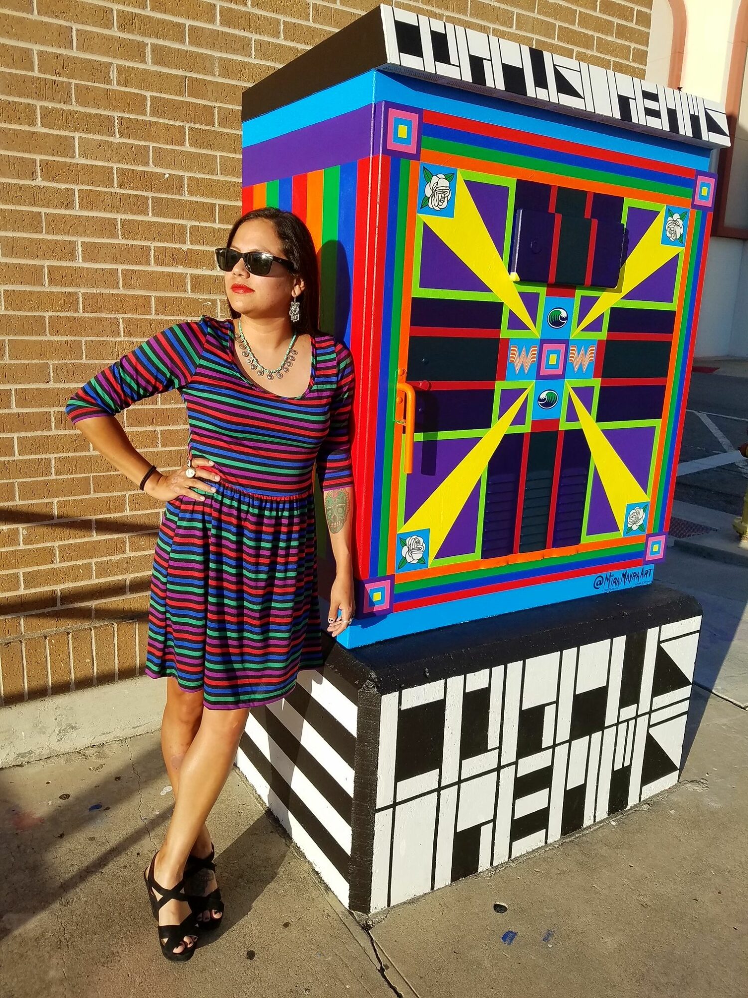 Woman in striped dress standing next to colorful painted box with geometric patterns, brick wall in background.