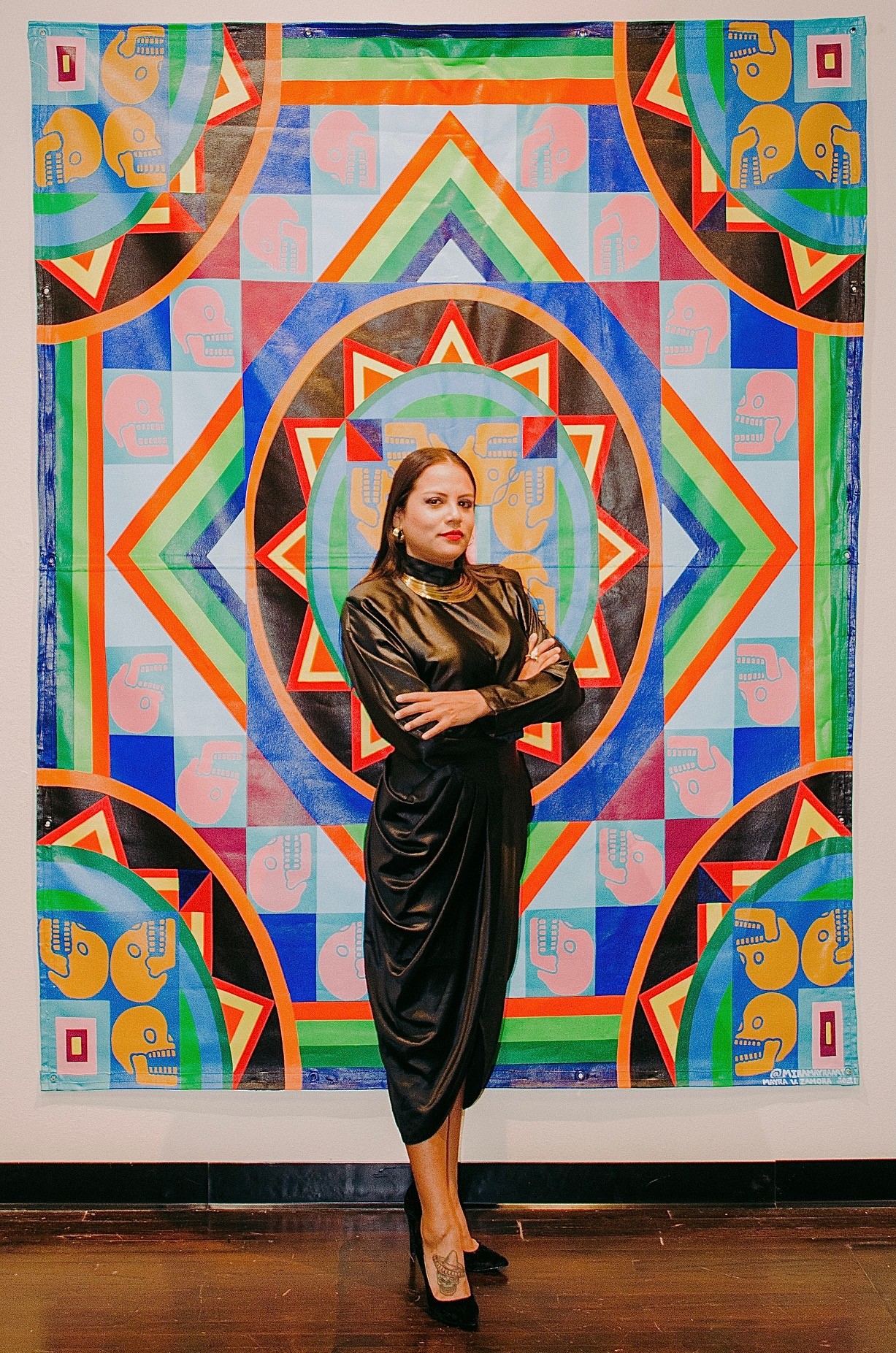 Woman in black dress standing with arms crossed in front of a colorful geometric mural.