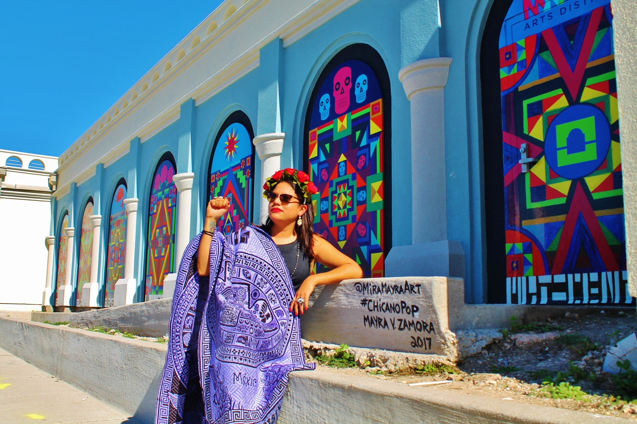 Woman with sunglasses and floral headband stands in front of colorful, patterned windows on a blue building.