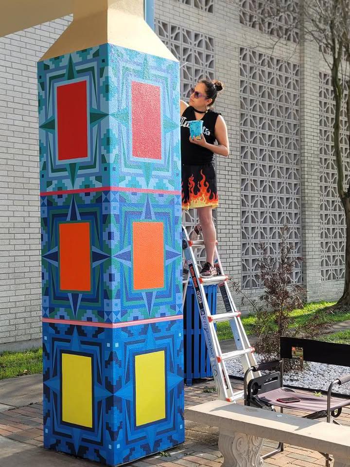 Person standing on a ladder painting a colorful, geometric mural on a tall column outdoors.