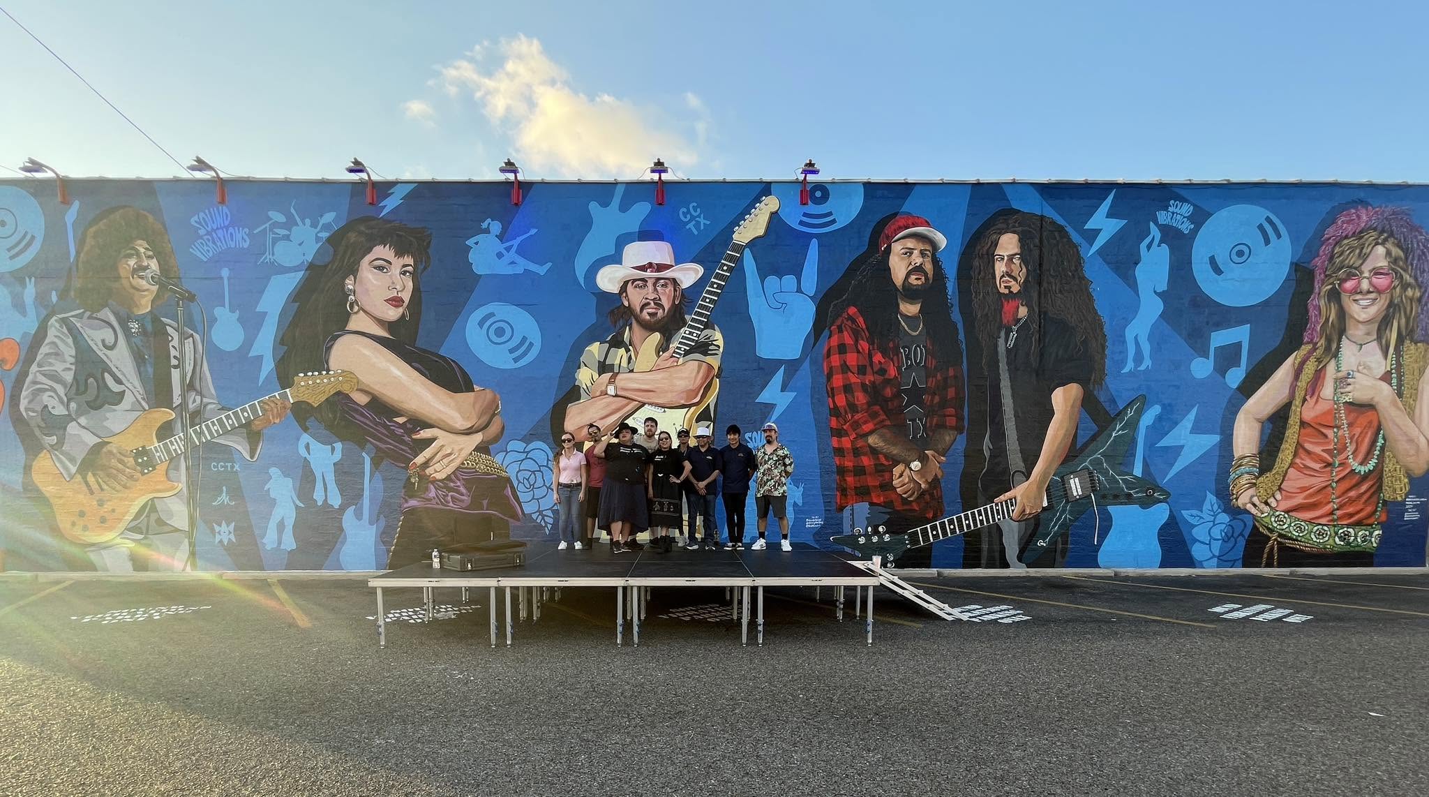 Large mural of six musicians playing instruments, with a blue background and various symbols, on a wall outdoors.