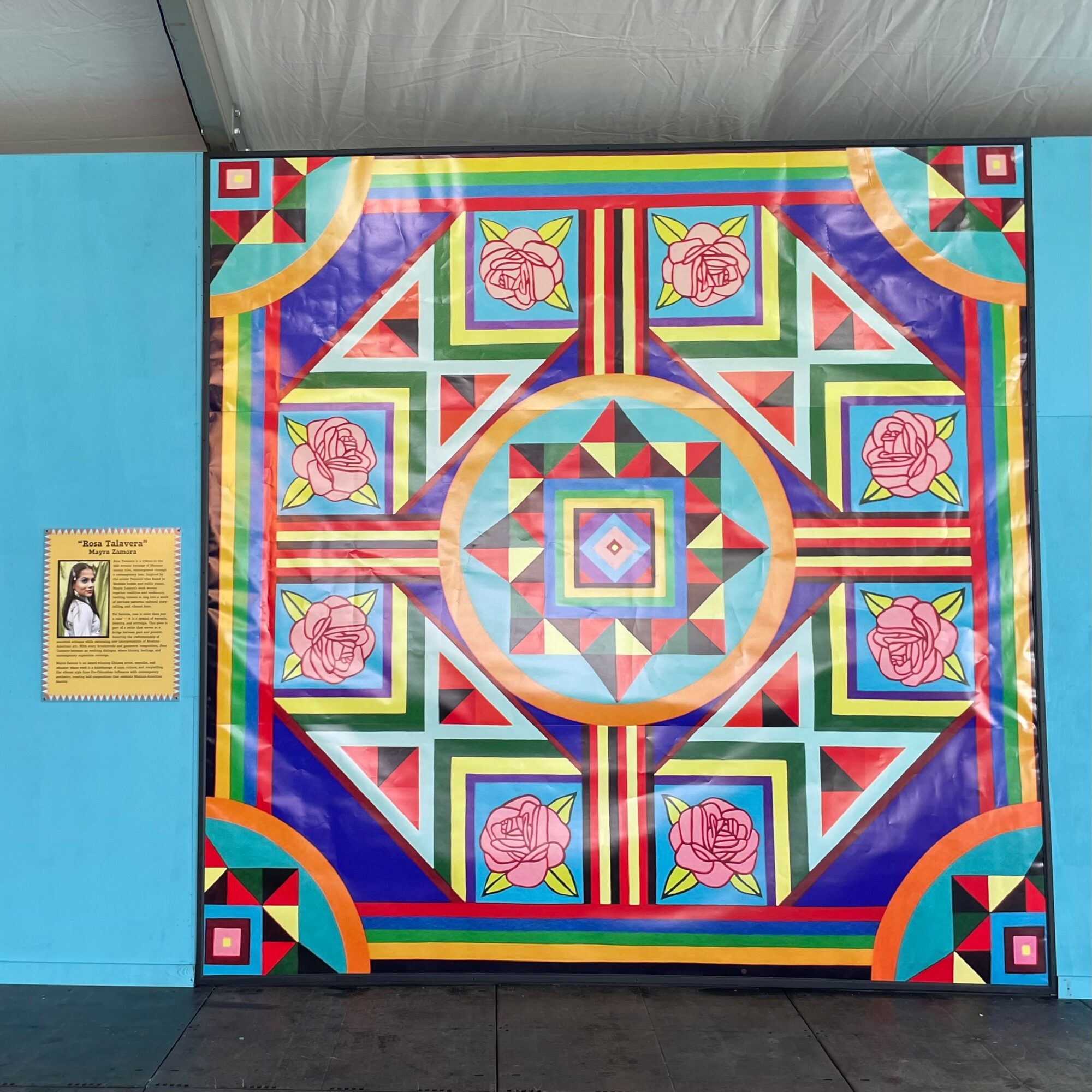 Colorful quilt with geometric patterns and pink roses, displayed on a wall with a blue background, and a small informational sign nearby.