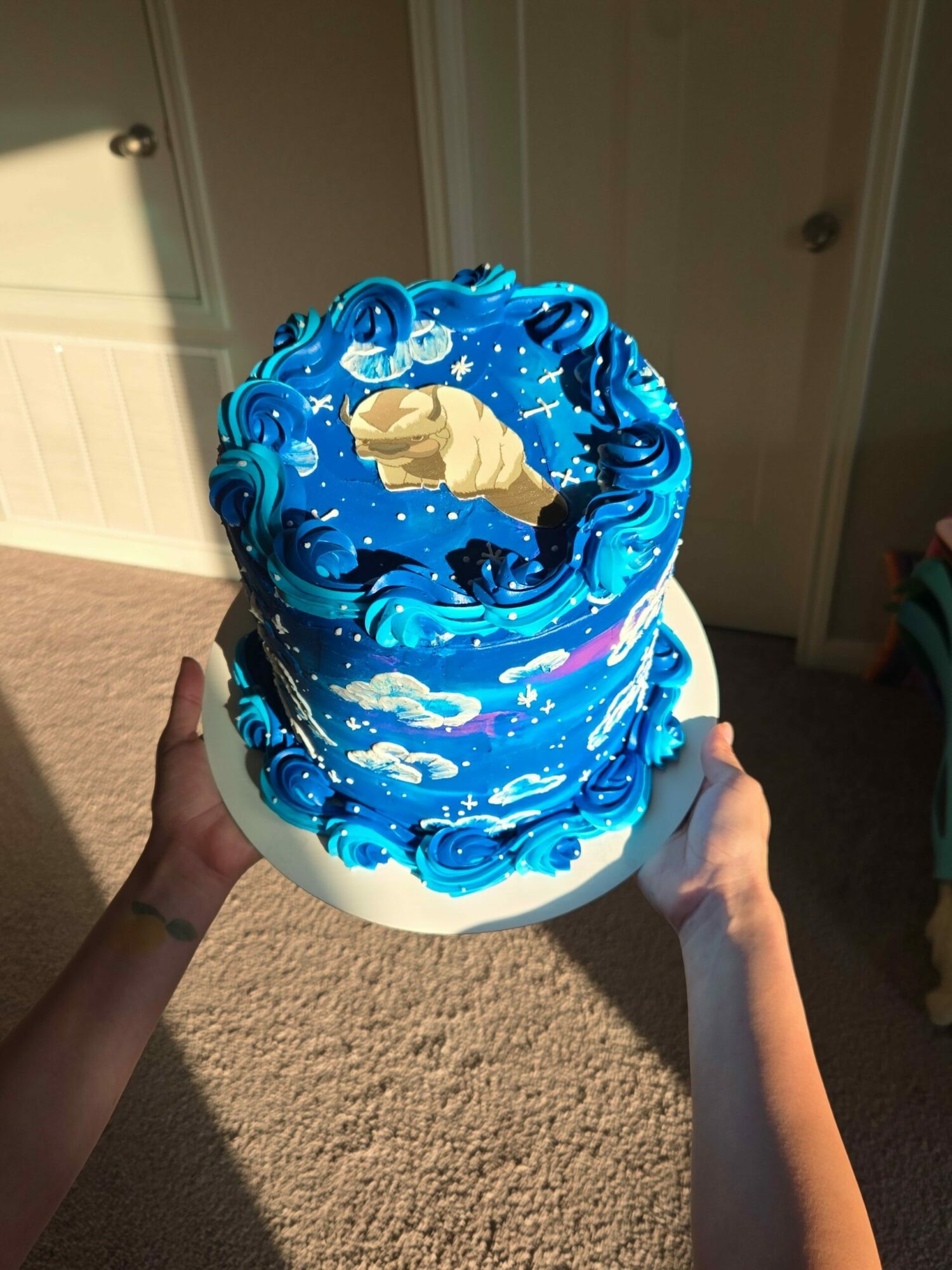 Person holding a blue, cloud-themed cake with decorative swirls and cloud designs, on a white cake stand.