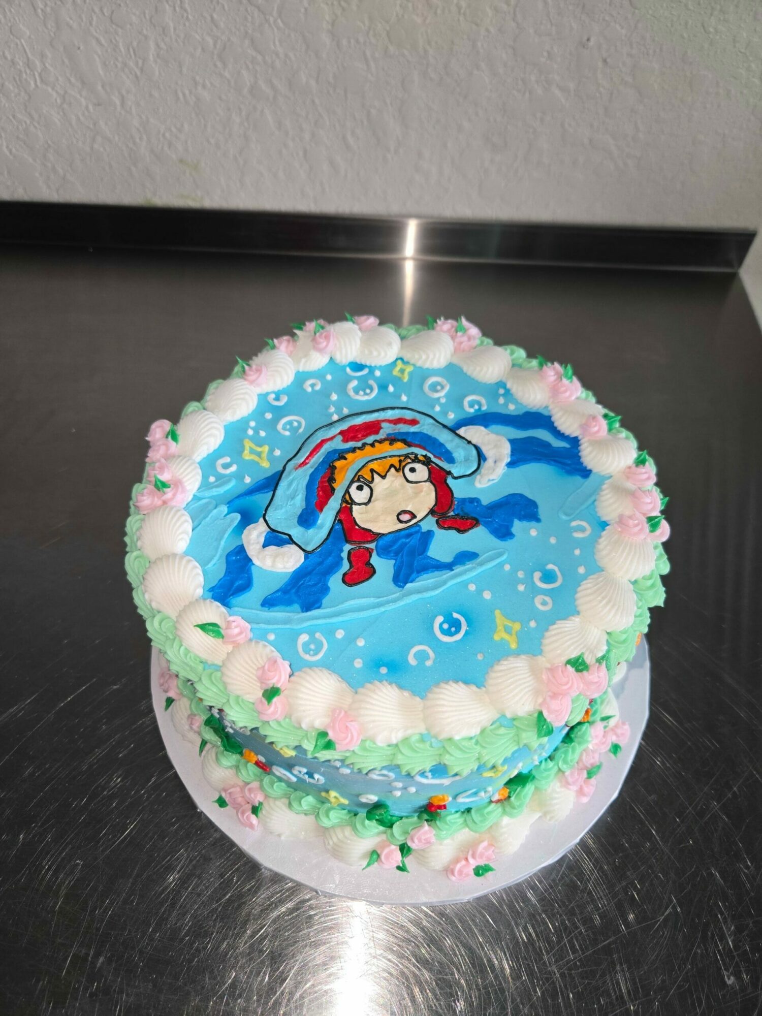 Round cake with a cartoon character swimming, decorated with pink, white, and green icing, on a silver surface.
