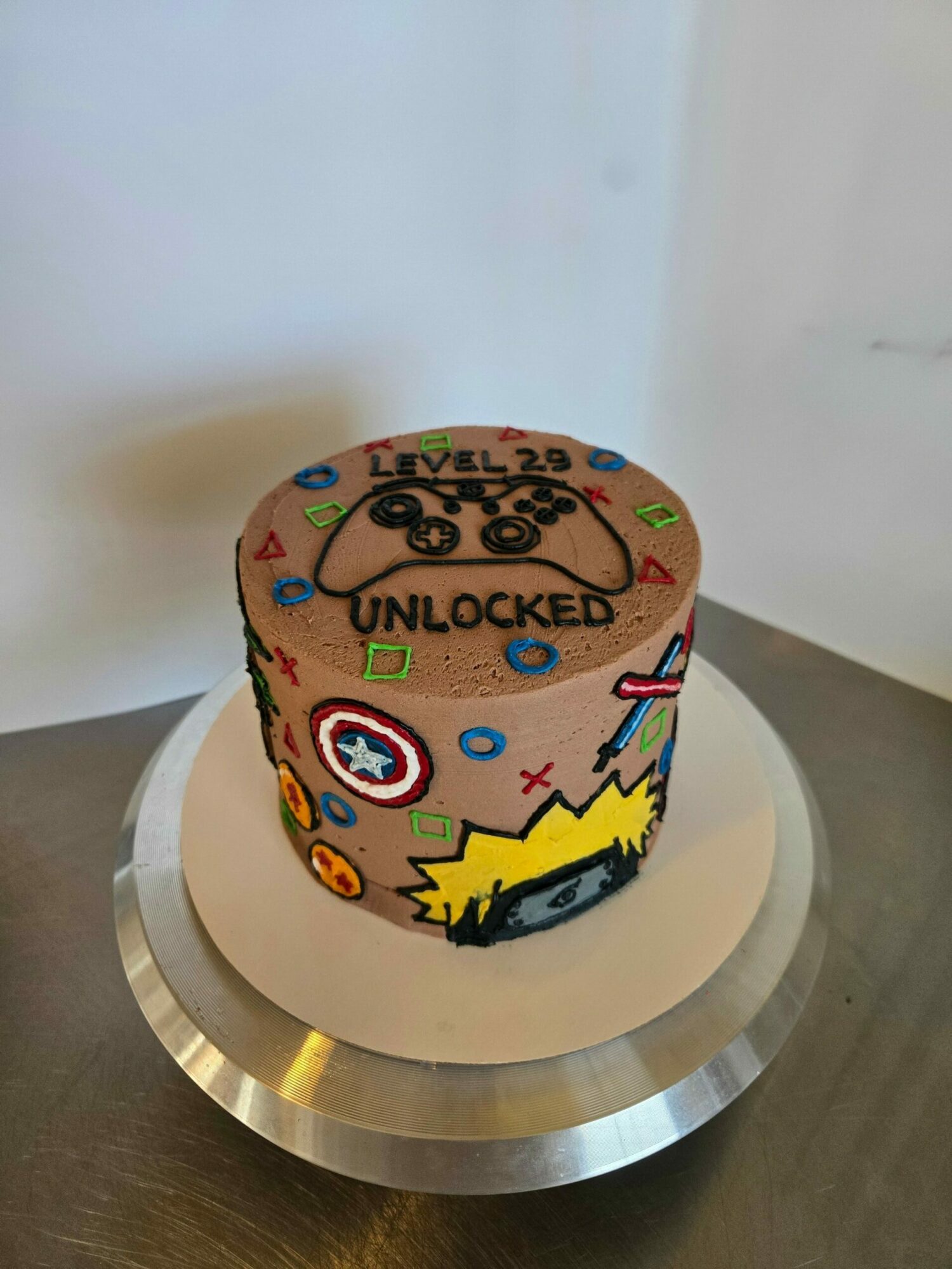 Cake decorated with superhero and gaming themes, including a black car, lightning bolt, and various symbols, on a white base.