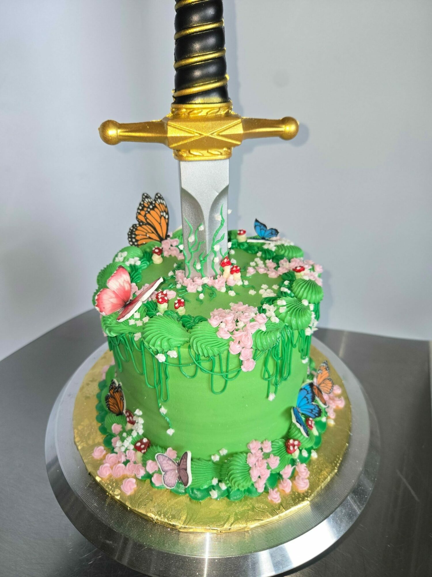 Cake decorated with green icing, flowers, and butterflies, with a sword-shaped candle on top, on a silver cake stand.