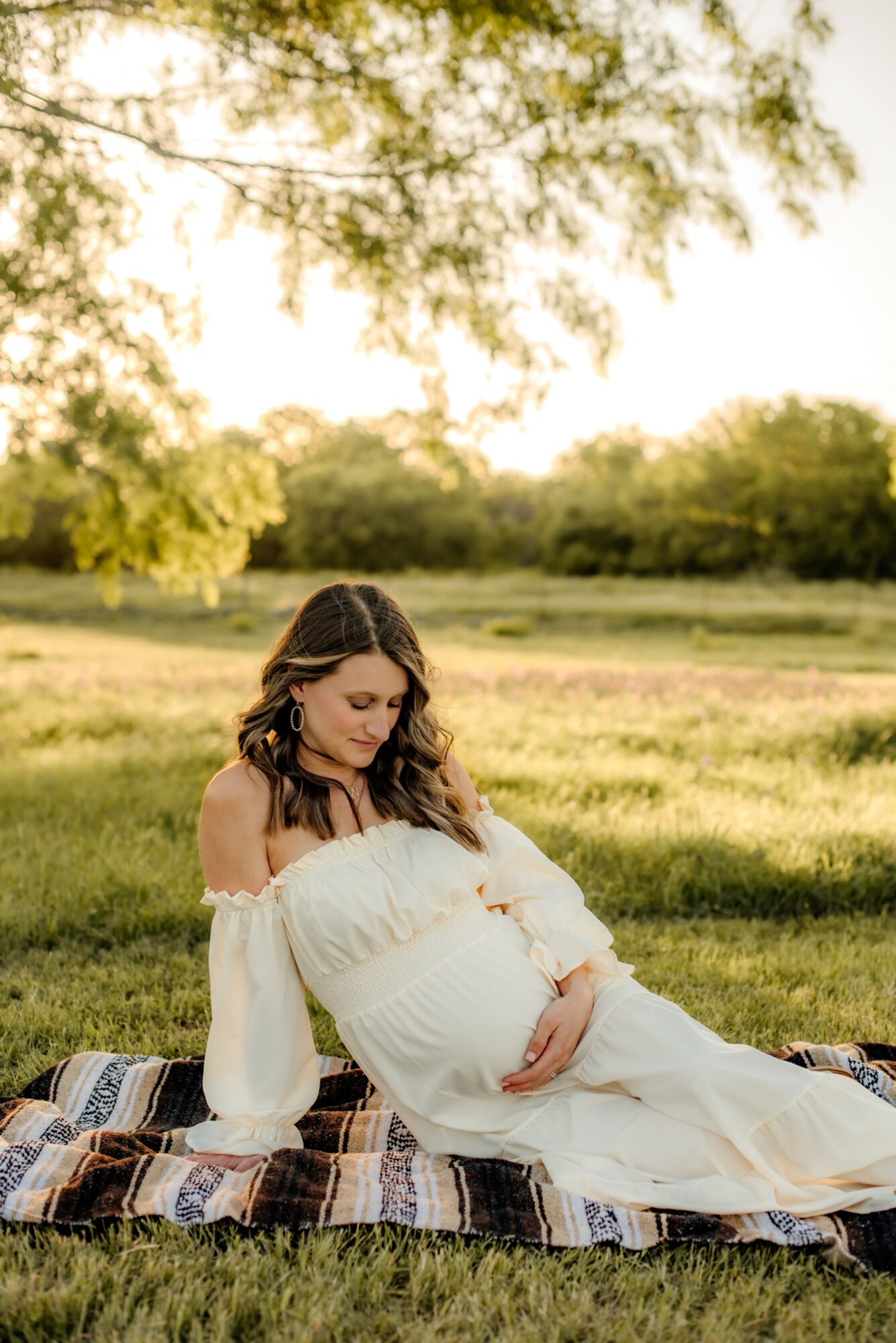 Pregnant woman sitting on a blanket outdoors, holding her belly, with trees and grass in the background, sunlight filtering through leaves.