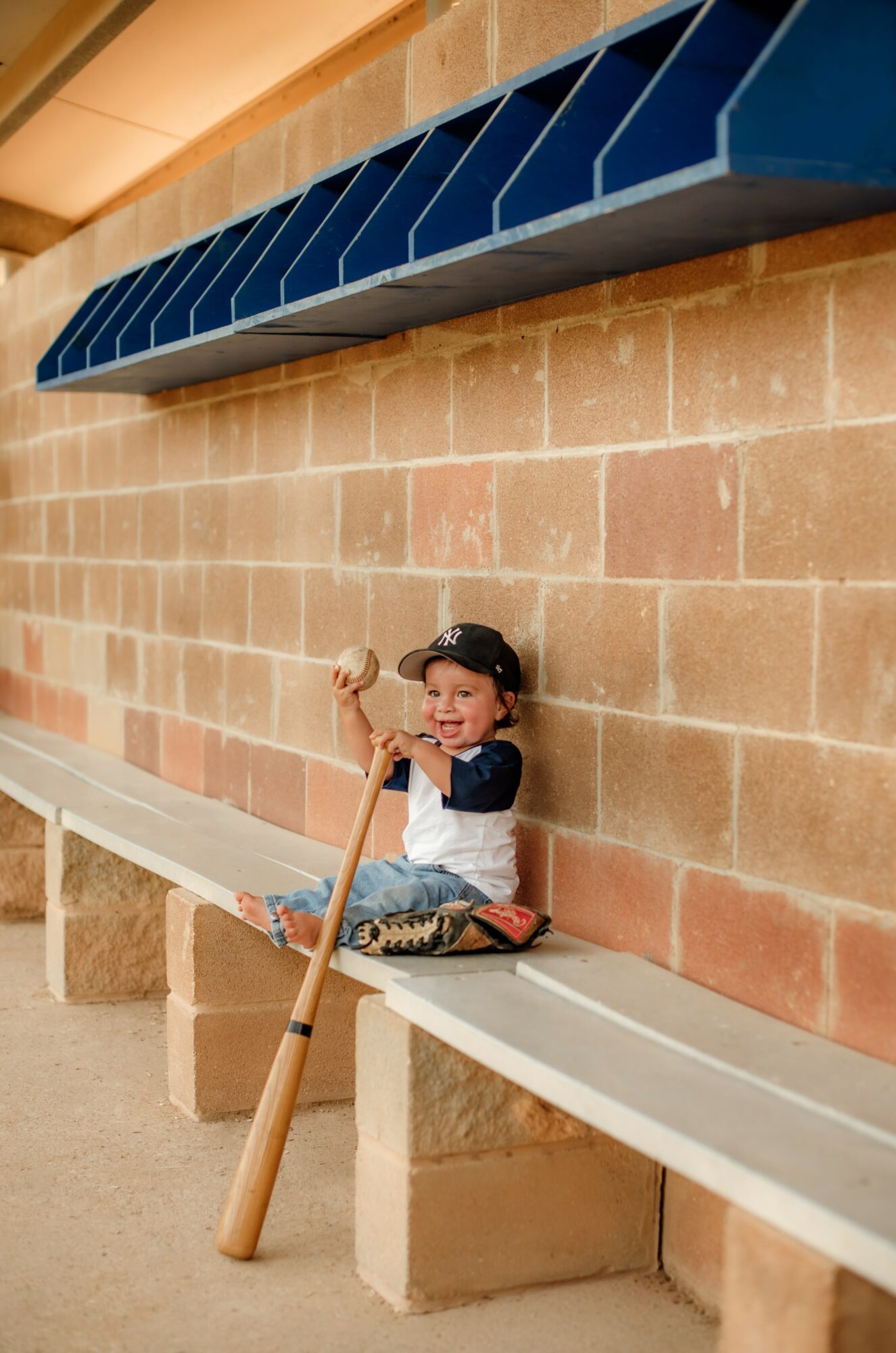 Child sitting on a white bench against a brick wall, holding a baseball bat, smiling, with a blue shelf above.