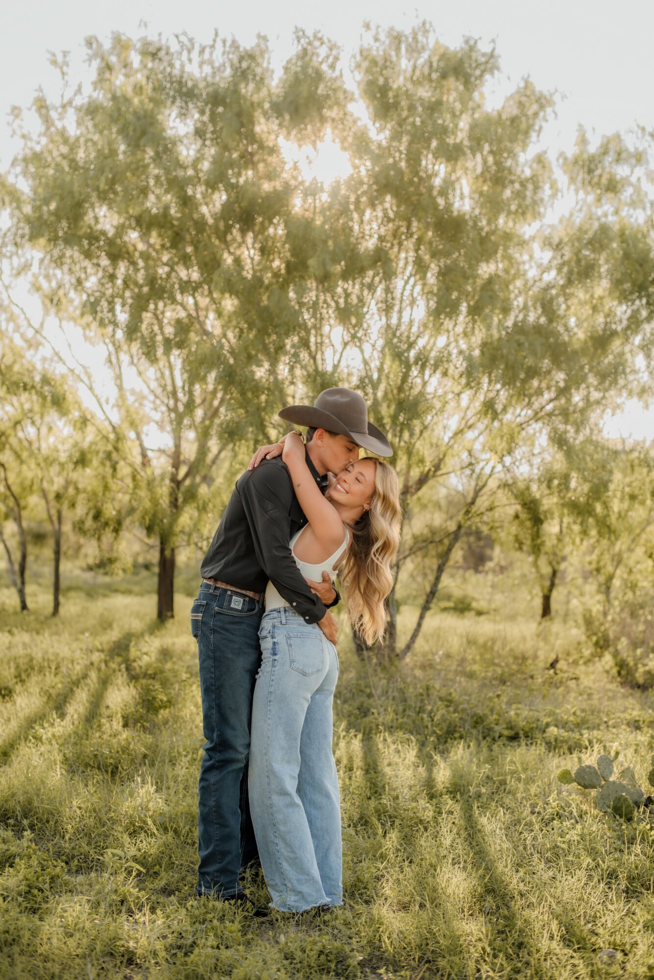 Two people hugging outdoors with trees and grass in the background, sunlight filtering through the leaves.