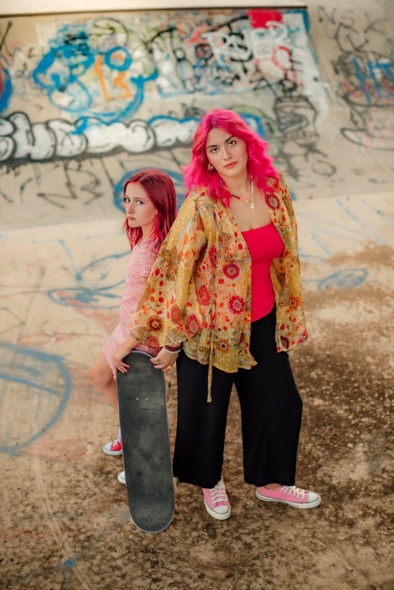 Two women with pink hair standing in front of a graffiti-covered wall, one holding a skateboard, outdoors.