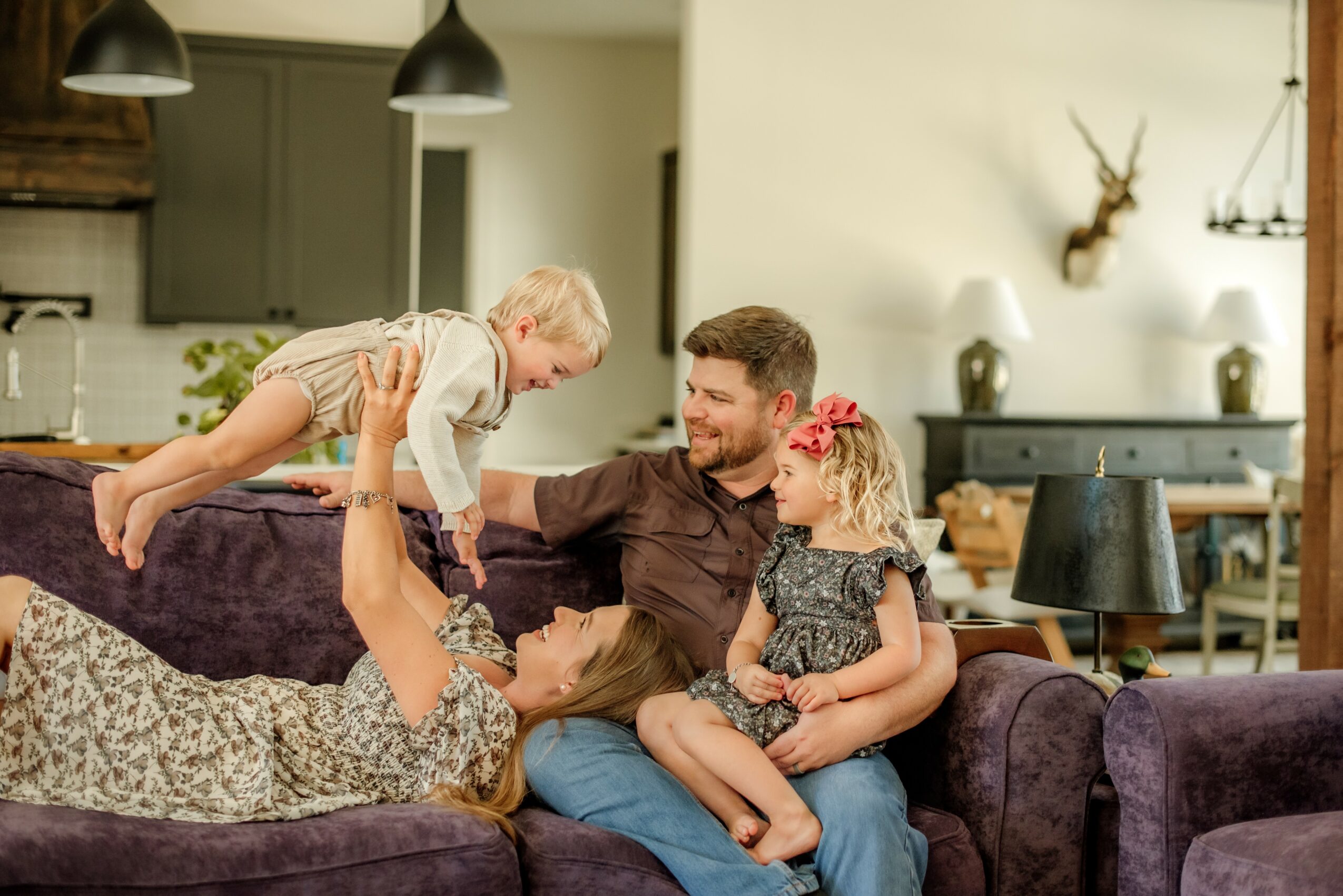 Family of four sitting on a sofa, playing and smiling together in a cozy living room, with a child being lifted by an adult.