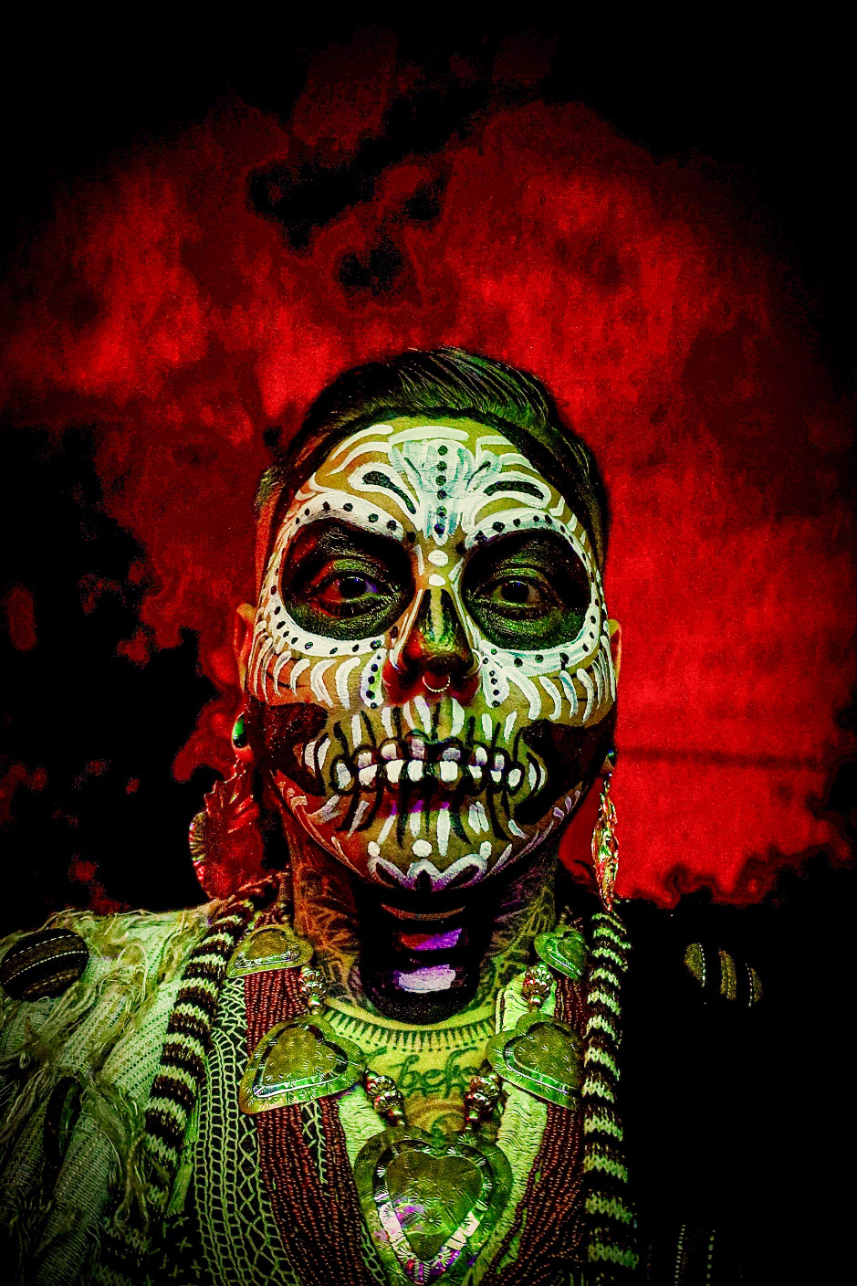 Person with face painted as a colorful skull, wearing jewelry, against a red and black background.
