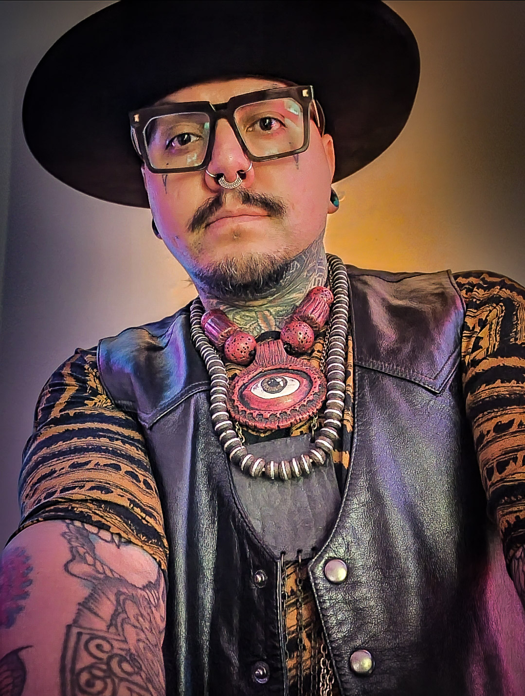 Person wearing glasses, a wide-brimmed hat, leather vest, and layered necklaces, with tattoos on arms, in colorful lighting.