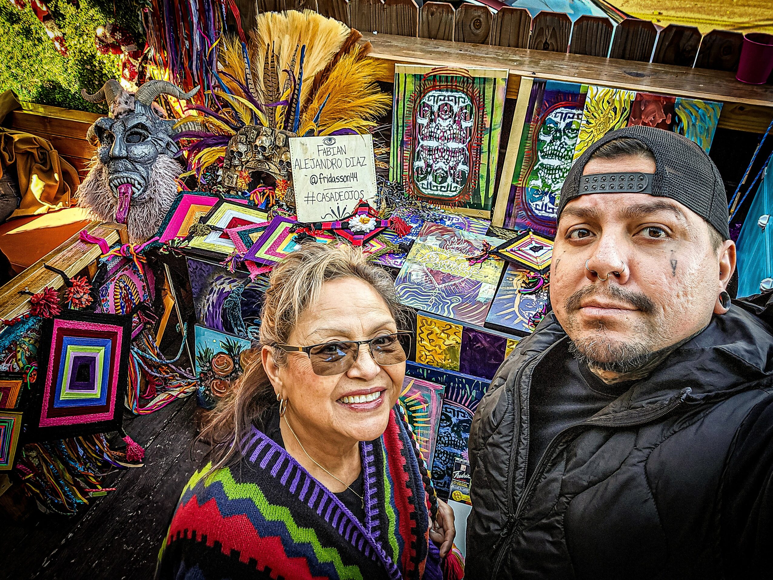 Two people smiling at a colorful market stall with textiles and crafts, including a wolf mask and vibrant woven items.