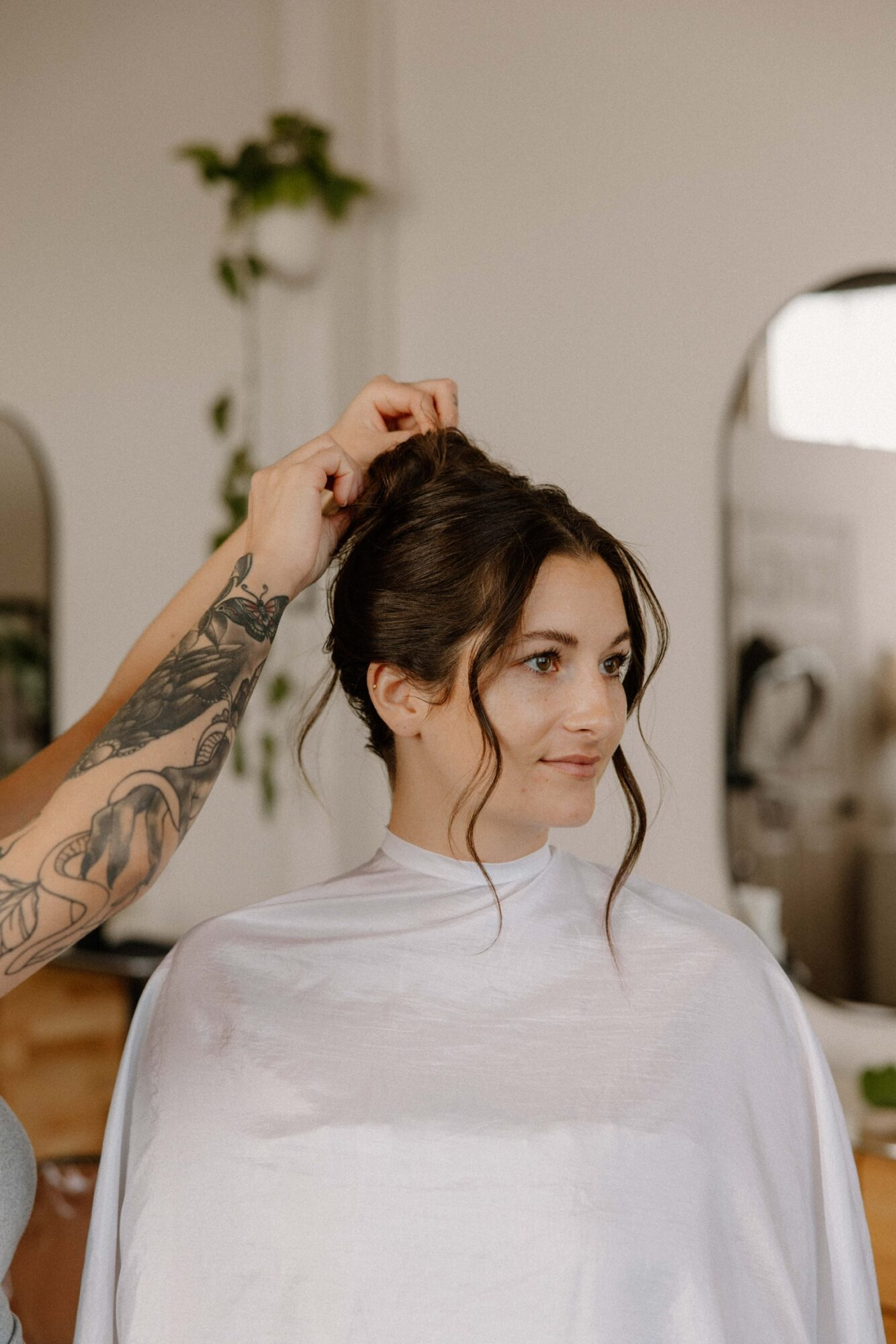 Woman with dark hair getting her hair styled by a person with tattoos in a salon, mirror in background.