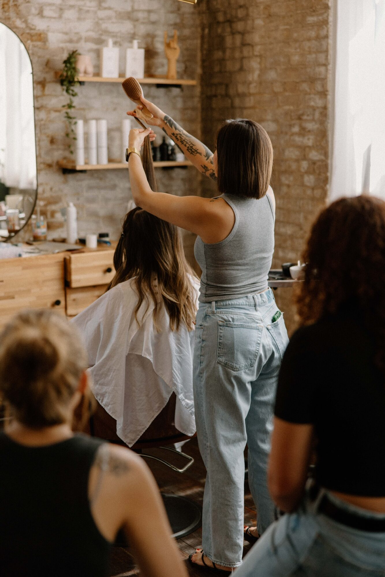 Woman demonstrating hairstyling to a group in a rustic room with brick walls and shelves.