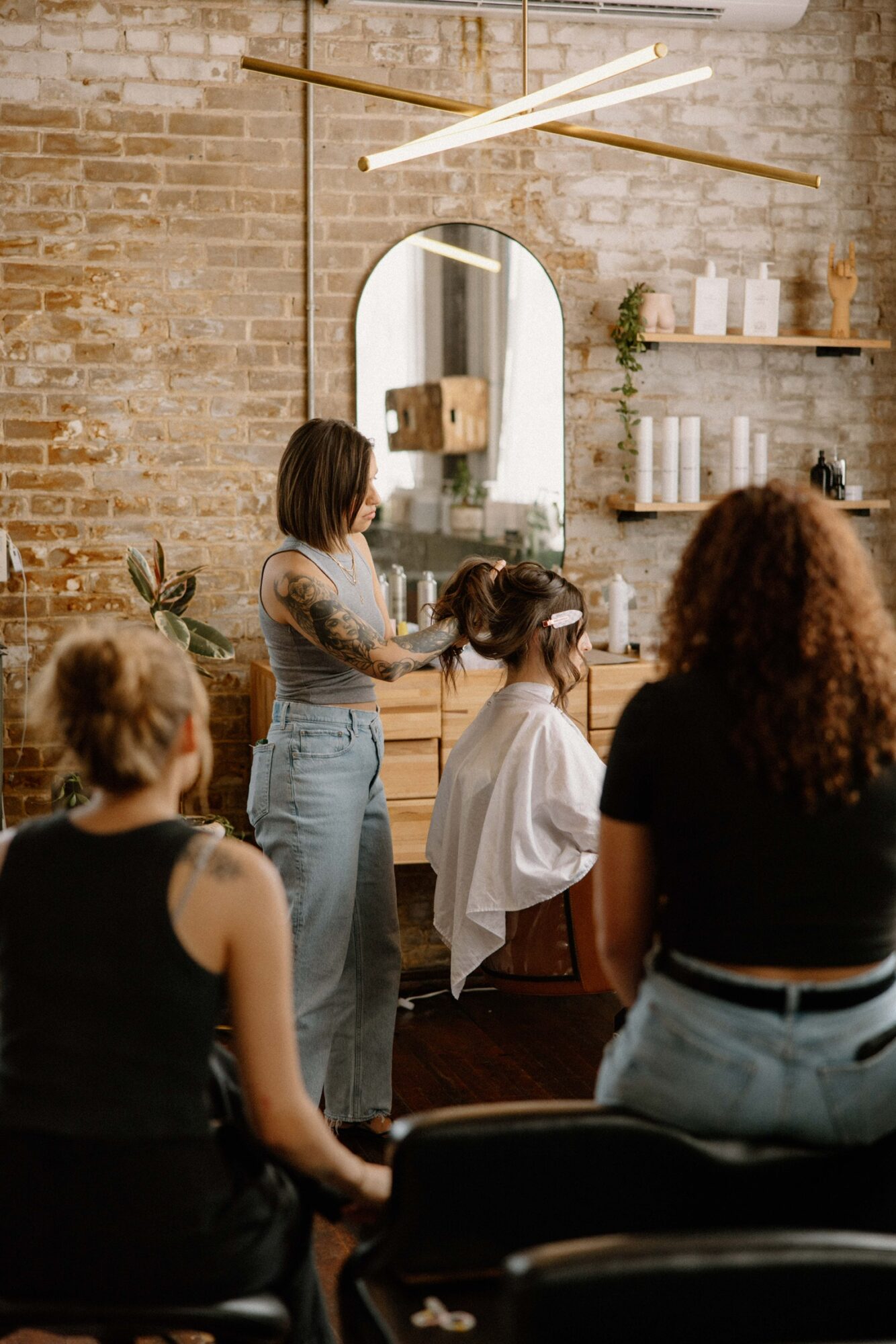 Group of women in a hair salon, one woman getting a haircut, mirror and shelves on brick wall, modern lighting, natural light