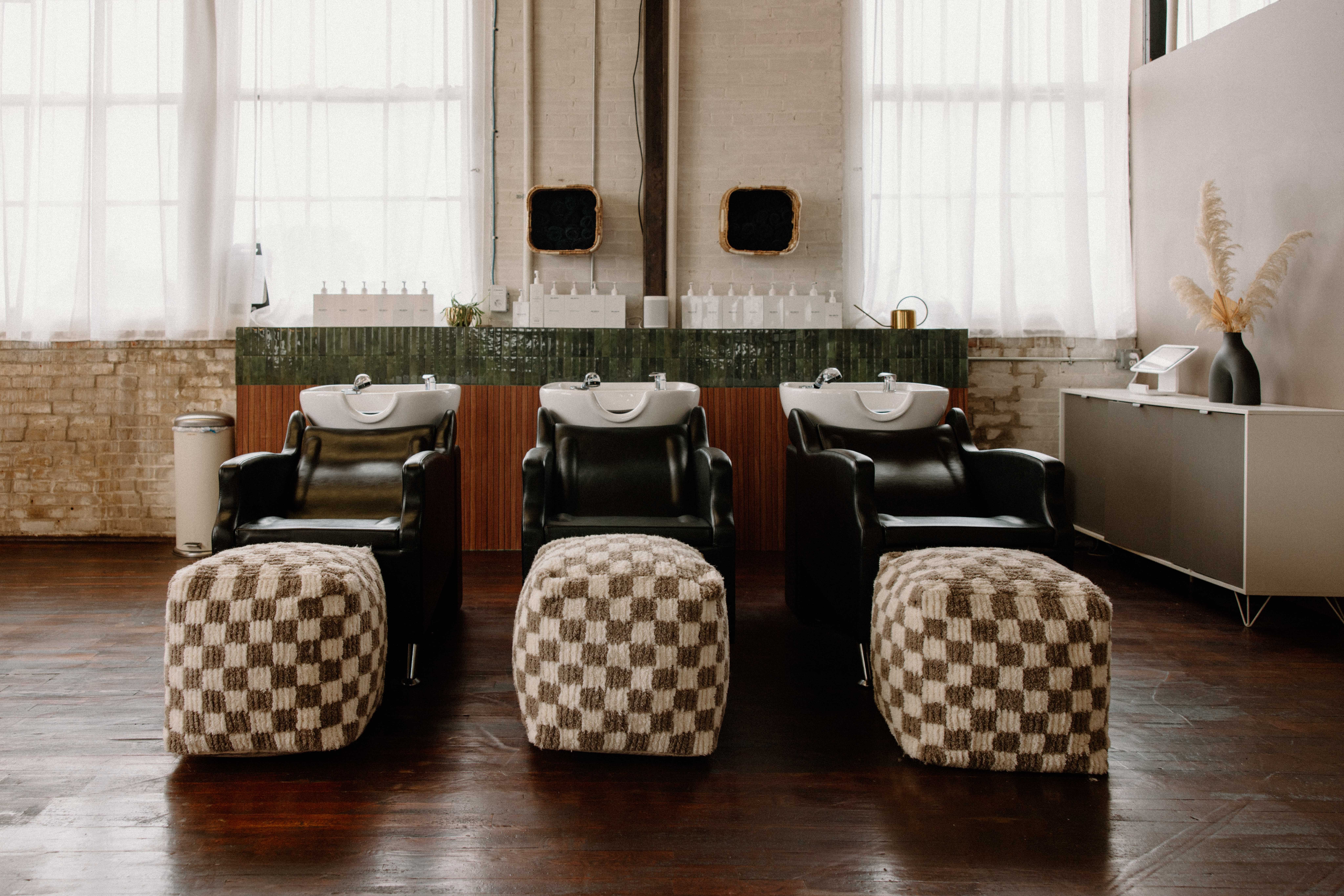 Three black salon chairs with checkered footstools in front, large windows with curtains, and a white cabinet with decorative items.