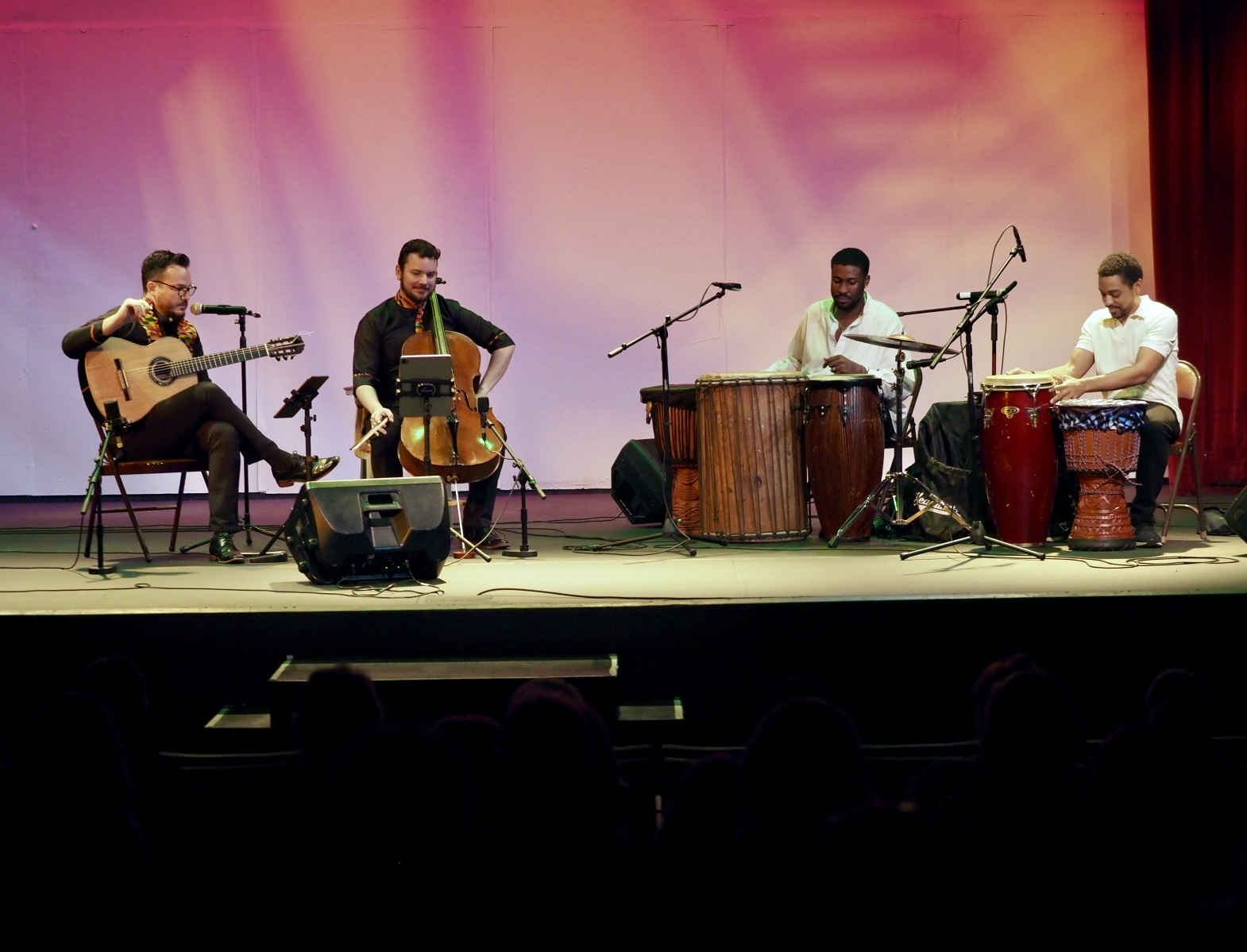 Four musicians perform on stage with instruments, colorful backdrop, and microphones, seated in a row with music stands.
