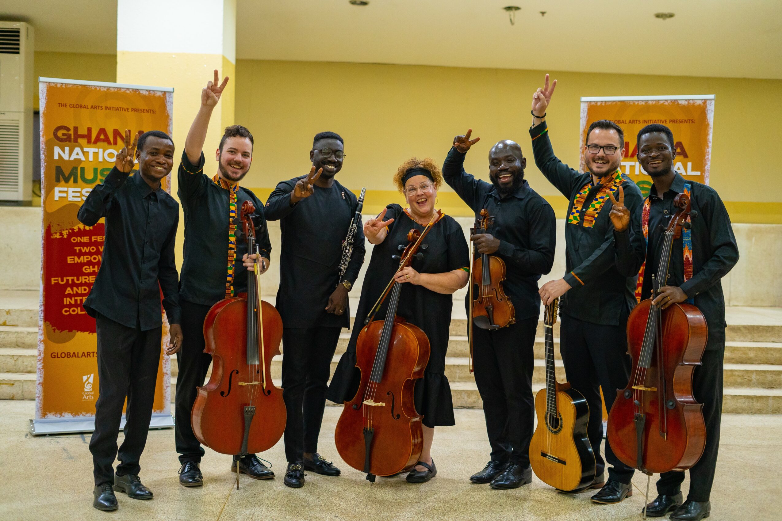 Group of seven musicians holding instruments, smiling, raising hands, standing indoors with banners in the background.