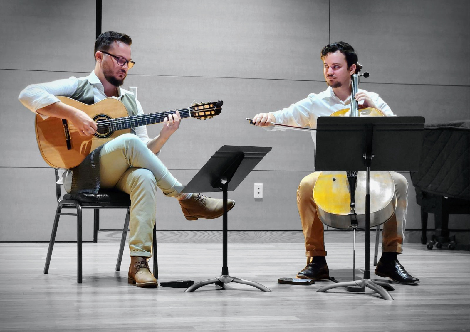 Two men playing guitars and a drum in a room with a gray wall, music stands, and a piano, seated and facing each other.