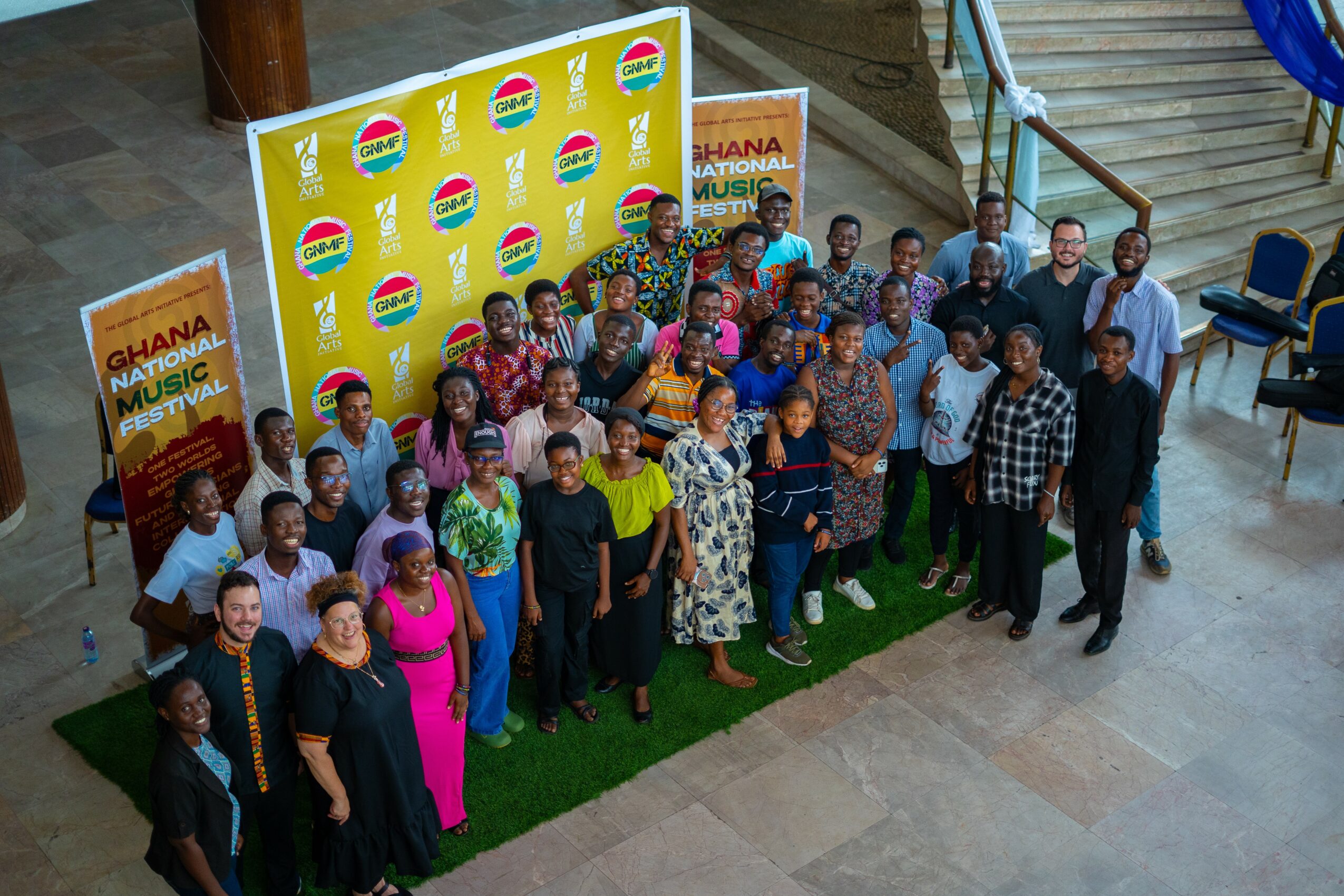 Group of people posing in front of a yellow backdrop with logos, some standing on green carpet, indoors.