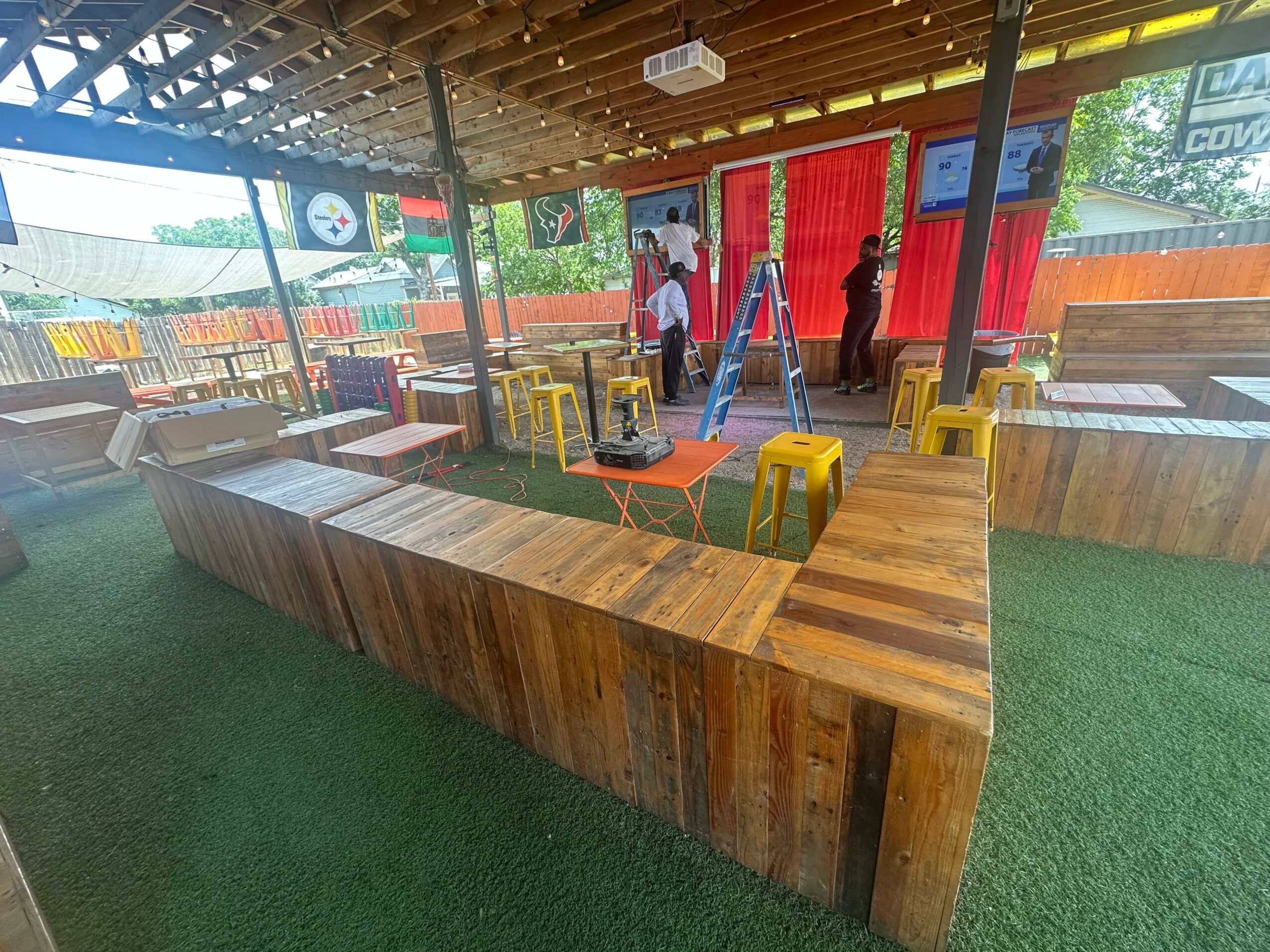 Indoor space with wooden seating, tables, and a ladder, with people near a red curtain and a projector on the ceiling.