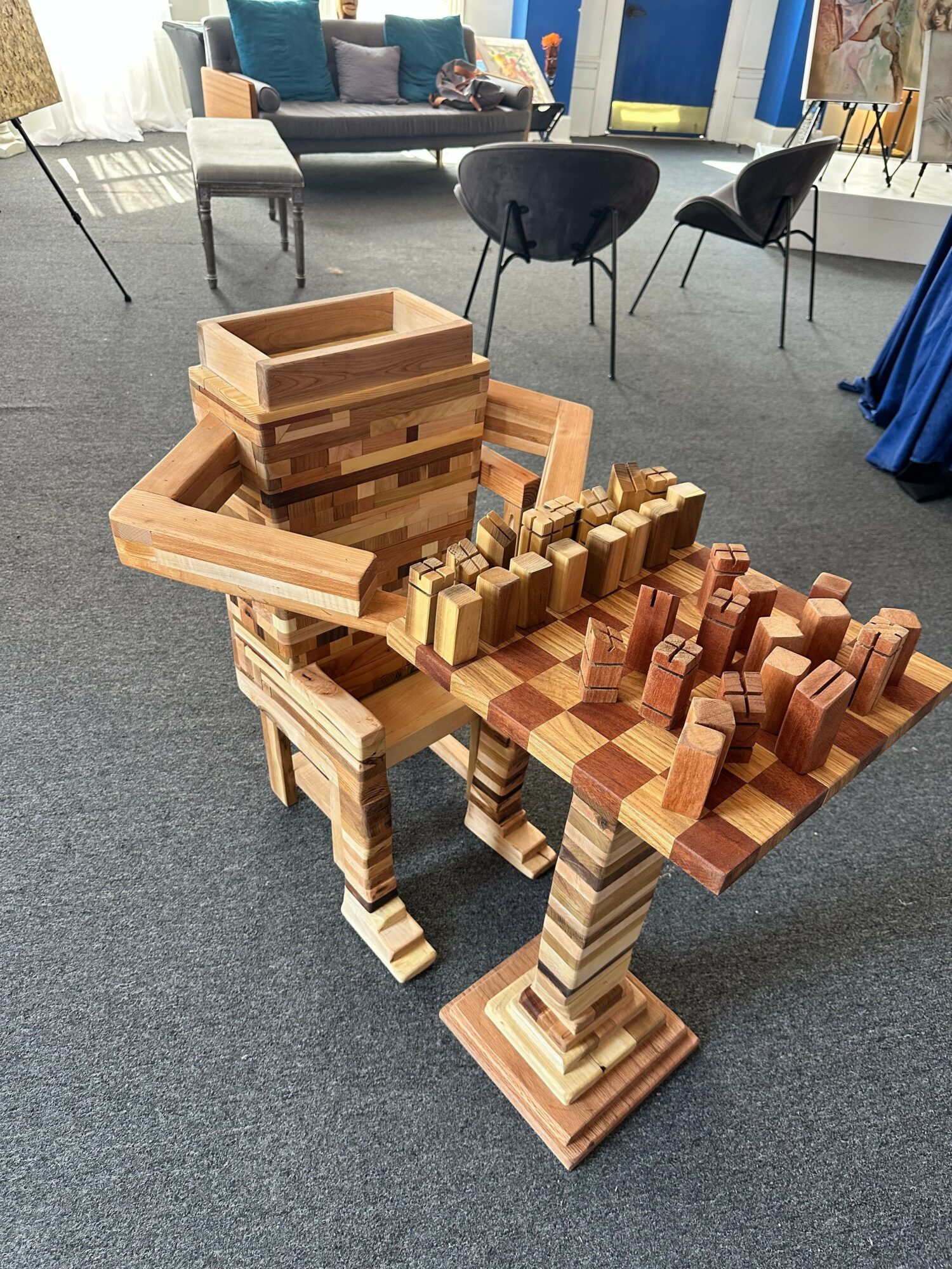Wooden chess pieces arranged on a table with chairs and a person in the background.
