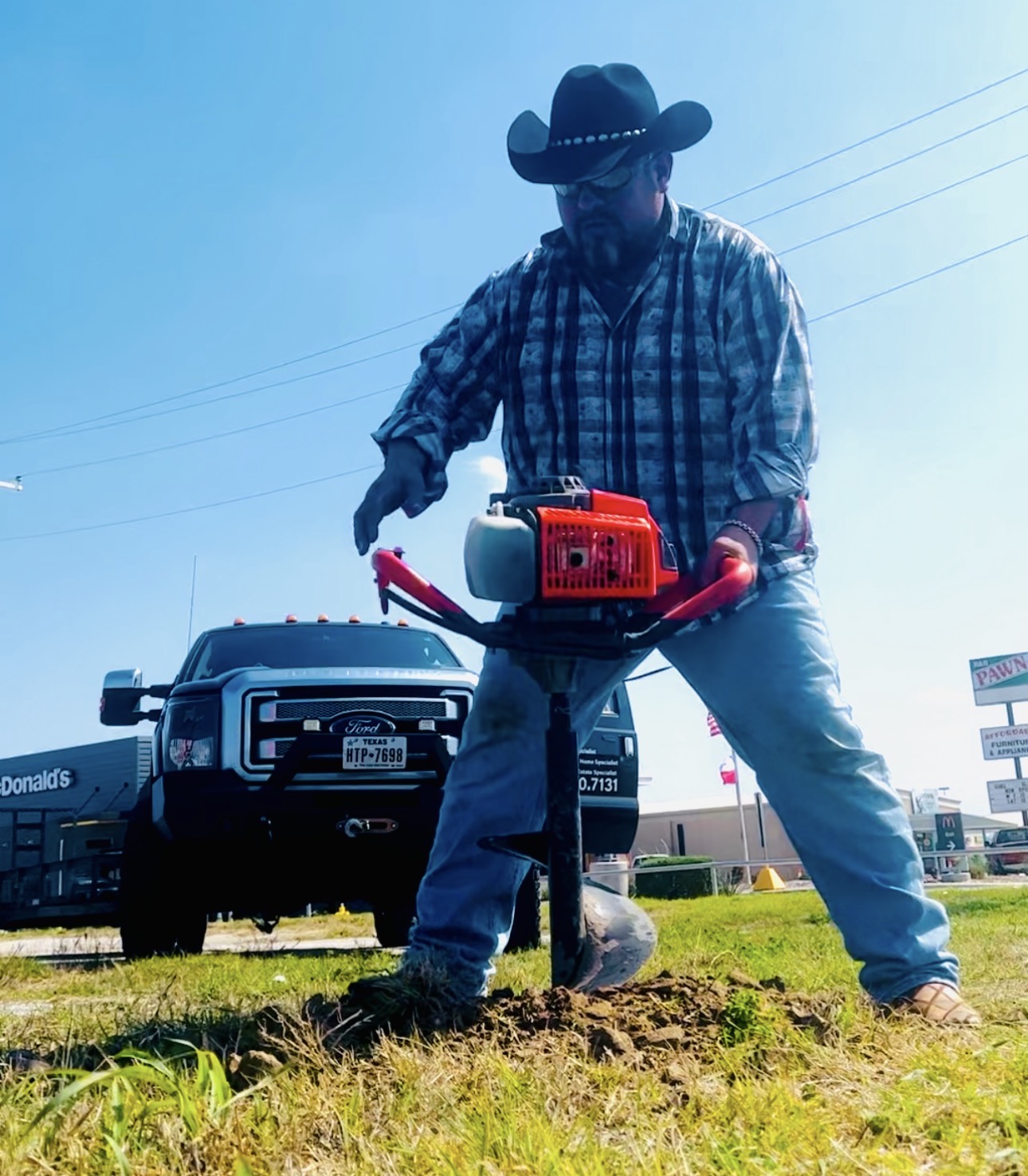 Person wearing a cowboy hat and plaid shirt using a red power tool on soil outdoors, with a vehicle and signs in background.