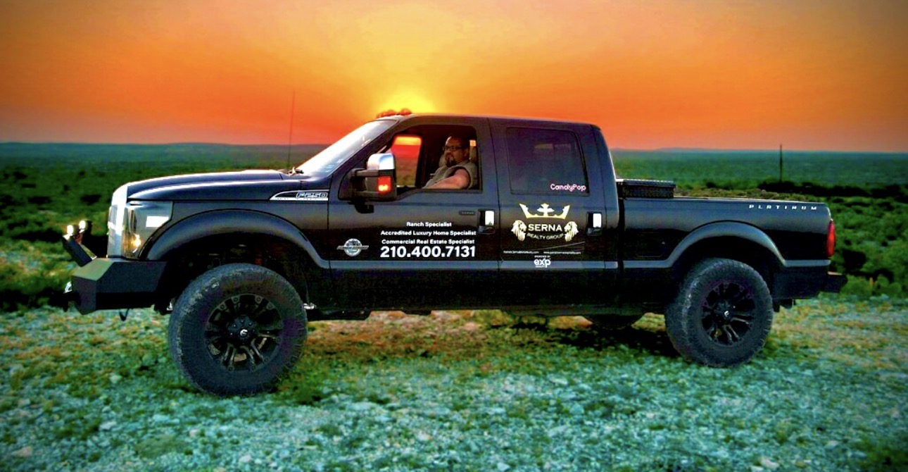 Black pickup truck parked on grass with sunset sky in background, driver visible inside, contact info on door, 20 words