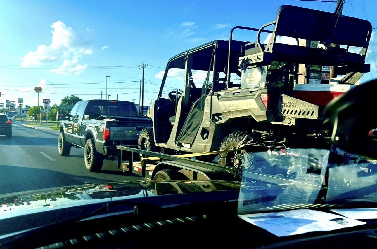 A vehicle with an open cargo area is on a flatbed truck, seen from inside a car on the road.